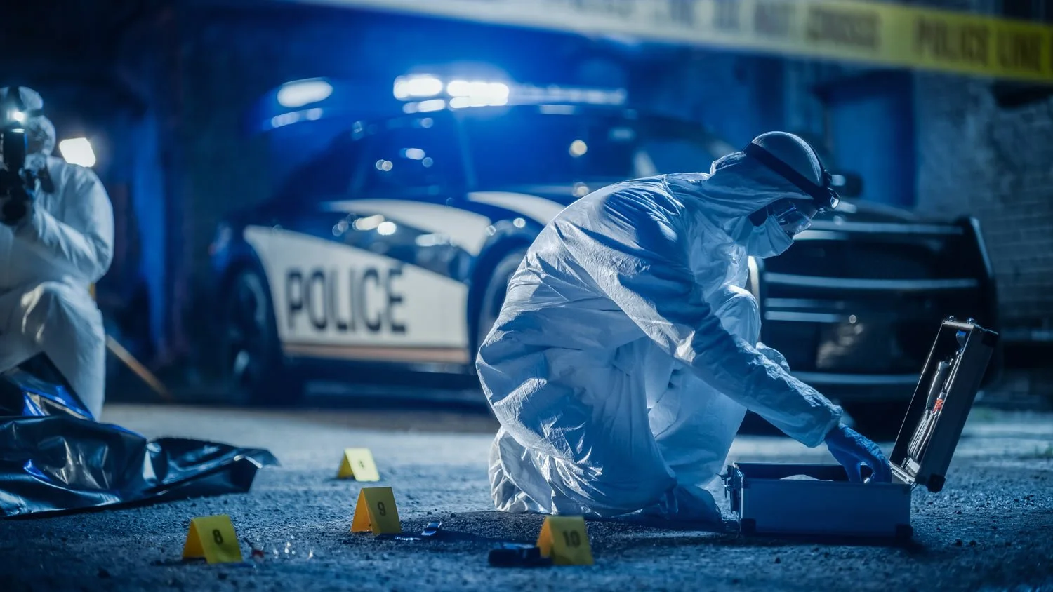 Forensic investigators in white protective suits collecting evidence on the street at night, police car with flashing lights in the background, yellow evidence markers on the ground.