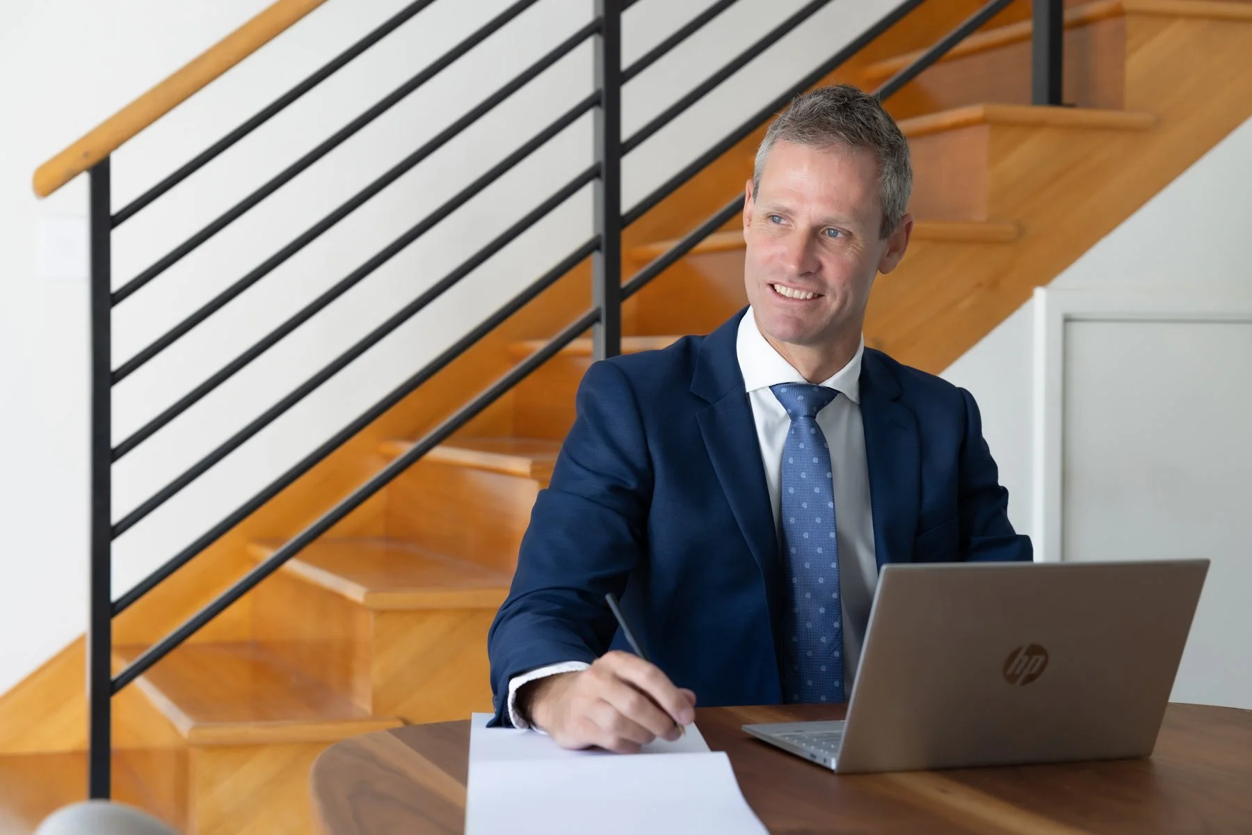 Dr Clint Hampson, Perth forensic expert and founder of Forensic Solutions WA, in a blue suit and tie, sitting at a desk with a laptop and writing on a notepad, in a room with a staircase in the background.