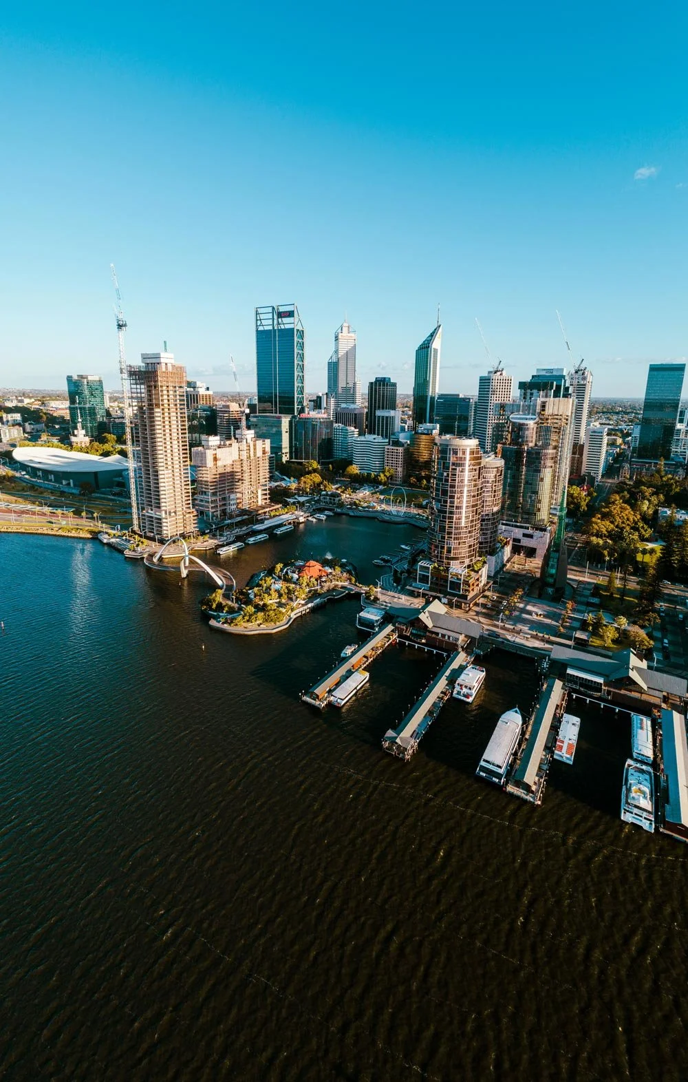 Aerial view of a Perth, Western Australia, city skyline along a harbor with tall skyscrapers, boats docked at piers, and a bridge with an arch over the water, under a clear blue sky.