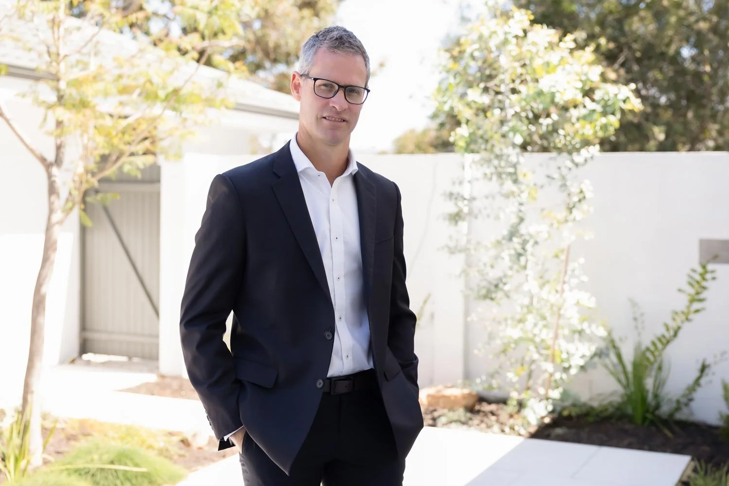 Dr Clint Hampson, Perth forensic expert and founder of Forensic Solutions WA, in a black suit and white shirt standing outdoors in front of a white fence and trees.