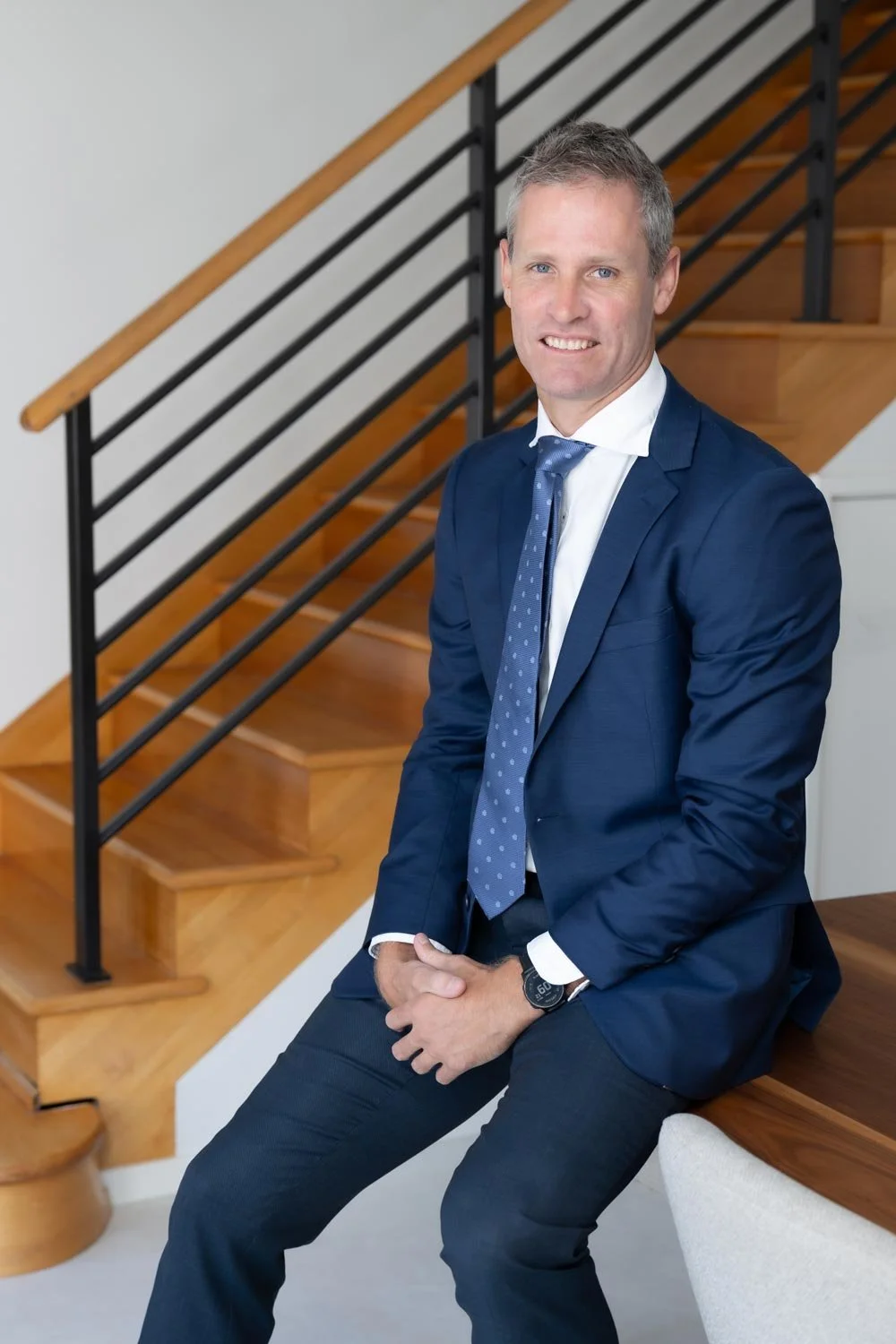 Dr Clint Hampson, Perth forensic expert and founder of Forensic Solutions WA, in a navy blue suit & tie, sitting on a white bench near a wooden staircase with black metal railing in a modern interior.