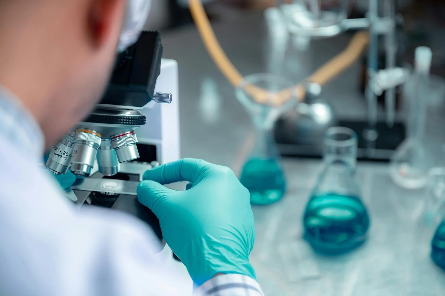 A Perth forensic scientist wearing gloves and examining a sample using a microscope in a laboratory setting with various glassware containing blue liquids, whilst meth testing Perth..