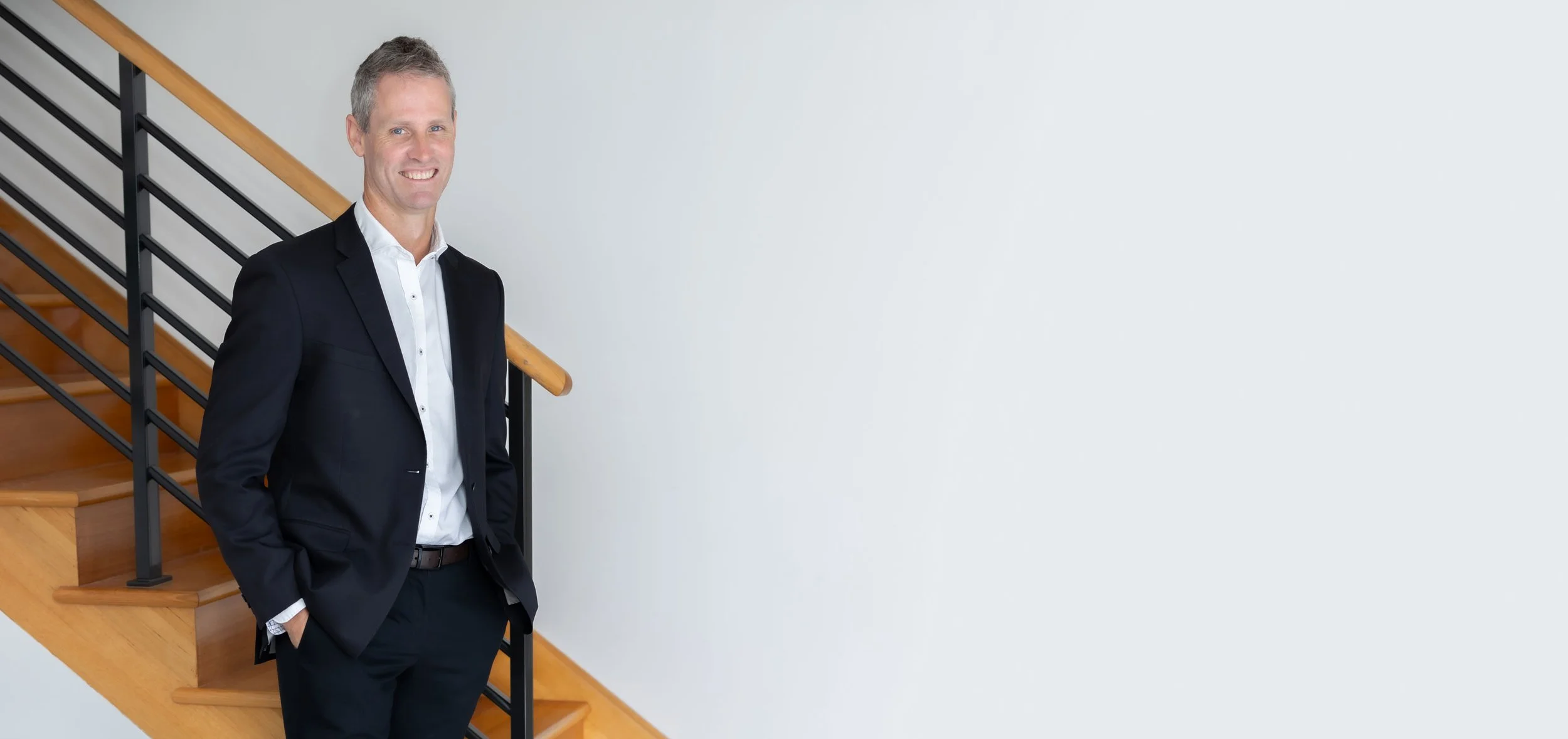 Perth forensic expert, Dr Clint Hampson, in a business suit standing on a wooden staircase indoors, smiling with hands in pockets.