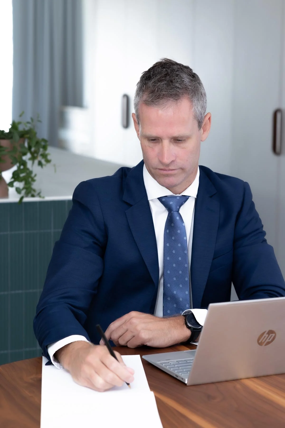 Dr Clint Hampson, founder of Forensic Solutions WA - offering expert forensic services in Perth, wearing a blue suit sitting at a wooden desk, writing on a piece of paper with a pen, in front of an open HP laptop.