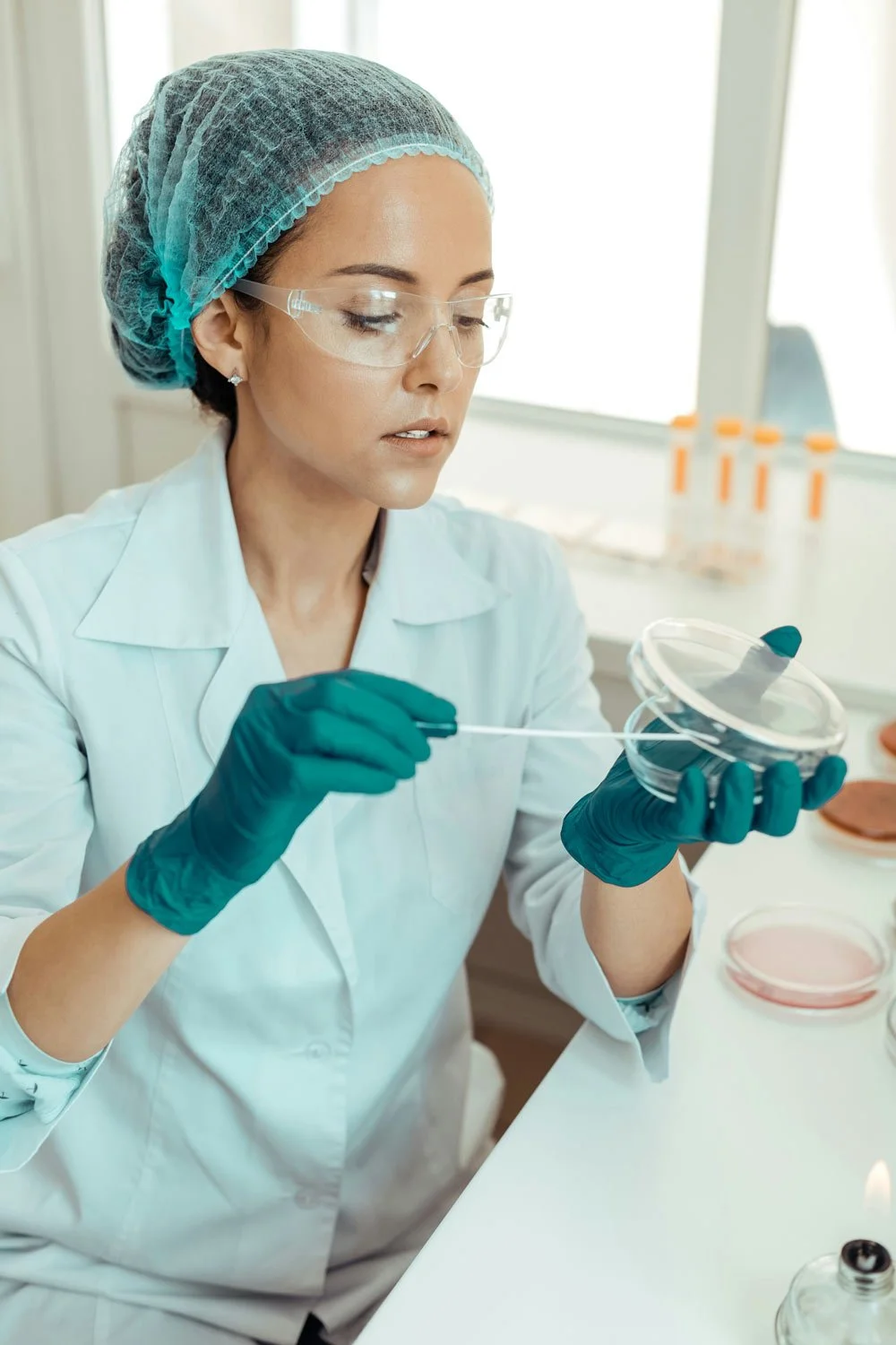 A female forensic scientist in safety goggles, gloves, and a hairnet is working with petri dishes in a laboratory.