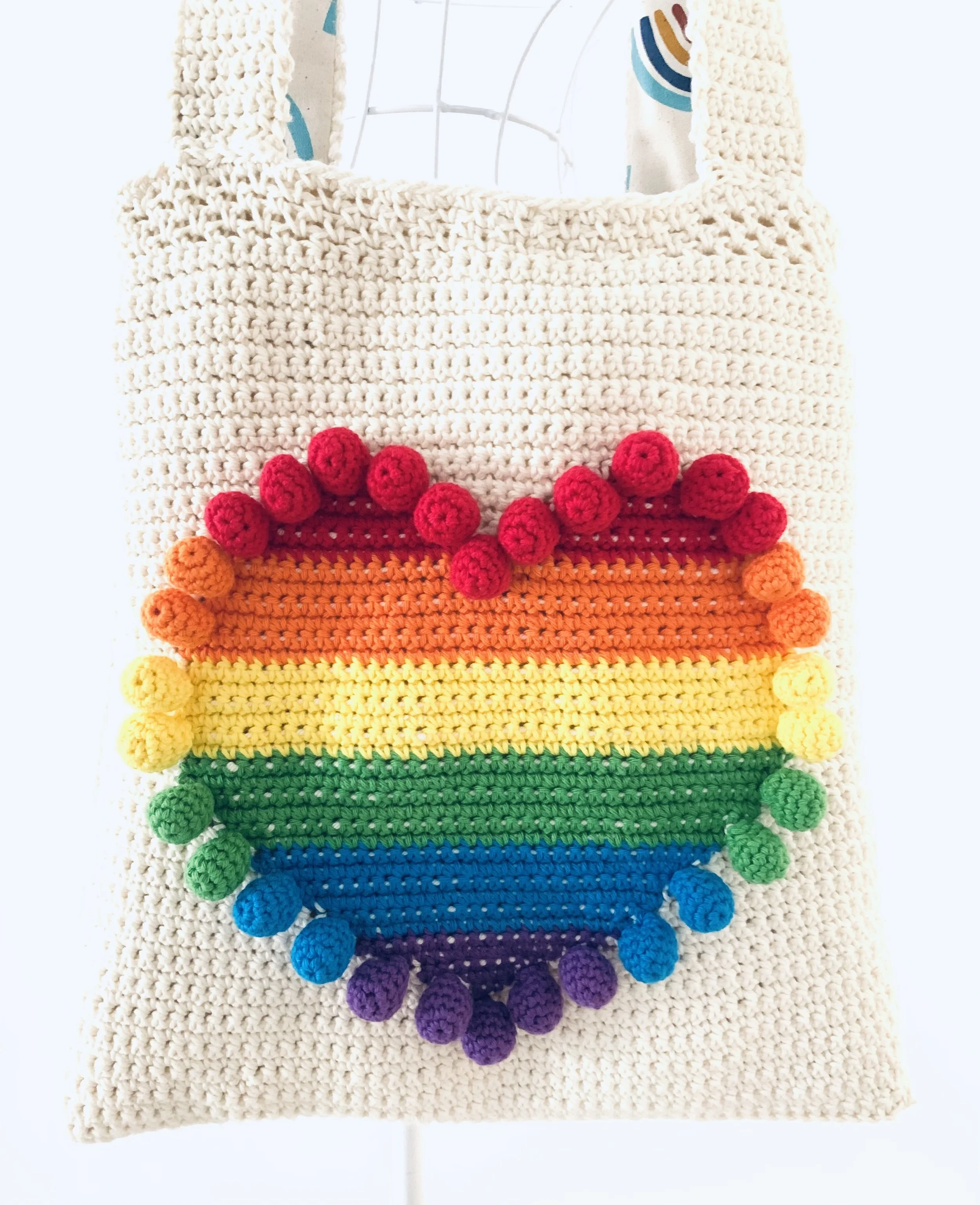 Handmade crochet tote bag with a rainbow-colored stripe pattern and multi-colored pom-pom trim.