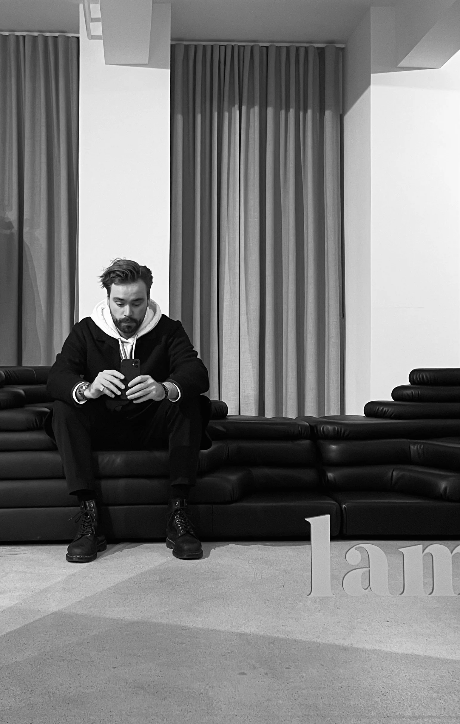 Junger Mann mit dunkler Kleidung und tätowierten Armen sitzt auf einer schwarzen Couch und schaut auf sein Smartphone. Im Hintergrund graue Gardinen und weiße Wände, im Vordergrund ein Teil eines weißen Buchstaben-Displays.