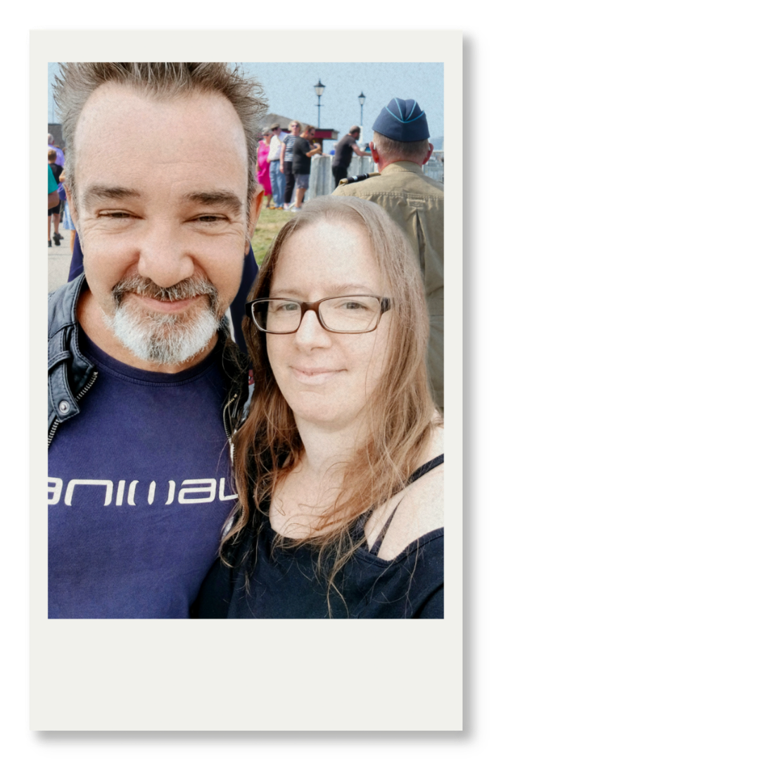 A man with short hair and beard, wearing a dark blue T-shirt and leather jacket, and a woman with long, reddish-brown hair and glasses, smiling at the camera at an outdoor event with people and lamp posts in the background.