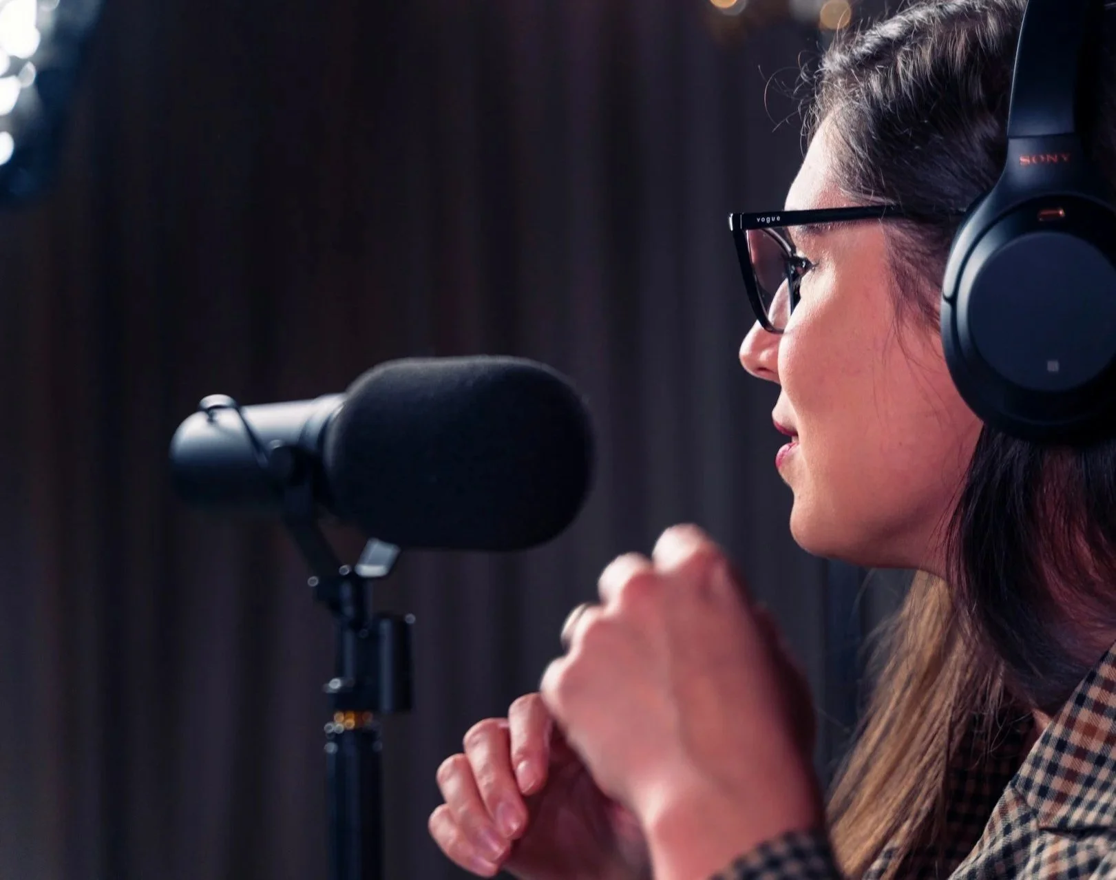 Naise henkilö puhuu mikrofonin ääreen radiostudioon, hänellä on kuulokkeet ja lasit.