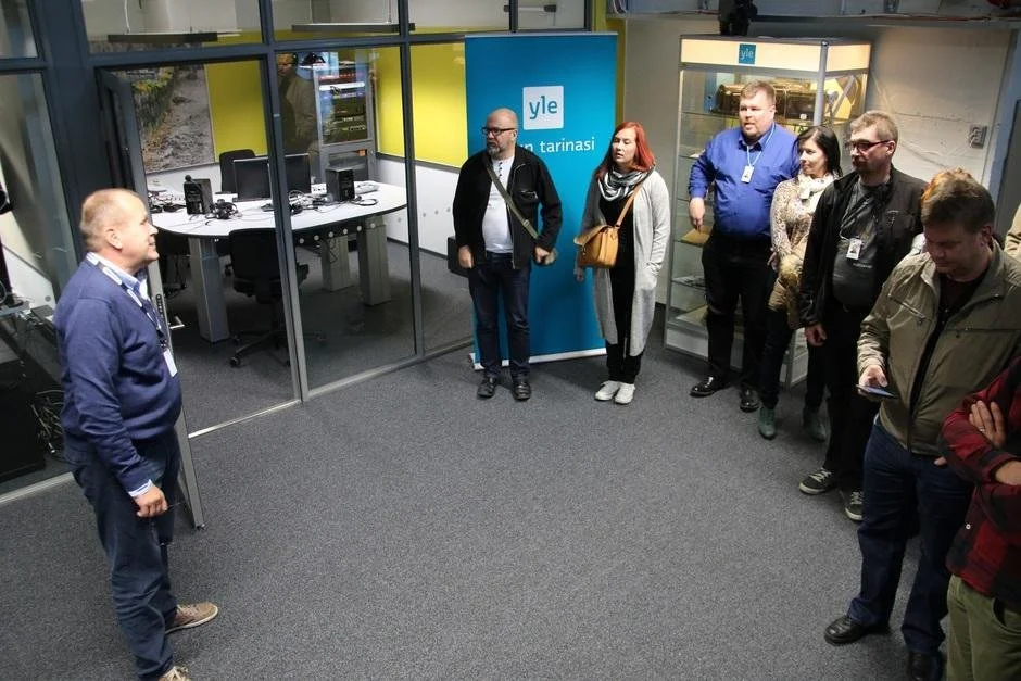 Ryhmä ihmisiä odottelee sisällä, keskustelutilassa, edessään esiintyjä, taustalla Yle-logo ja toimistotilaa.