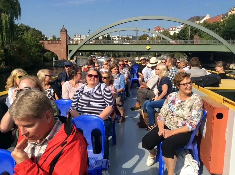 Ihmiset istuvat kannella laivalla matkalla sen ollessa joen päällä. Taustalla silta ja kaupungin rakennuksia.