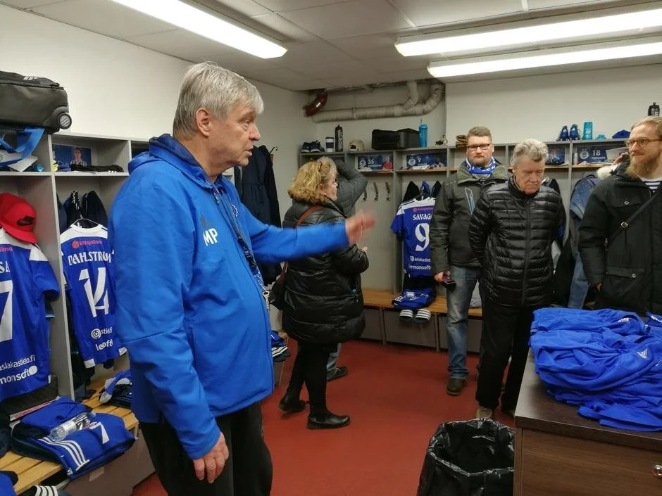 Ryhmän ihmiset pukuhuoneessa, joissa on jalkapallovarusteita ja -paitoja.