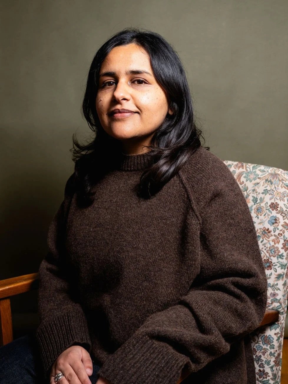 A brown woman with dark long hair and a brown knit sweater sitting on a floral-patterned chair against a plain dark green wall.