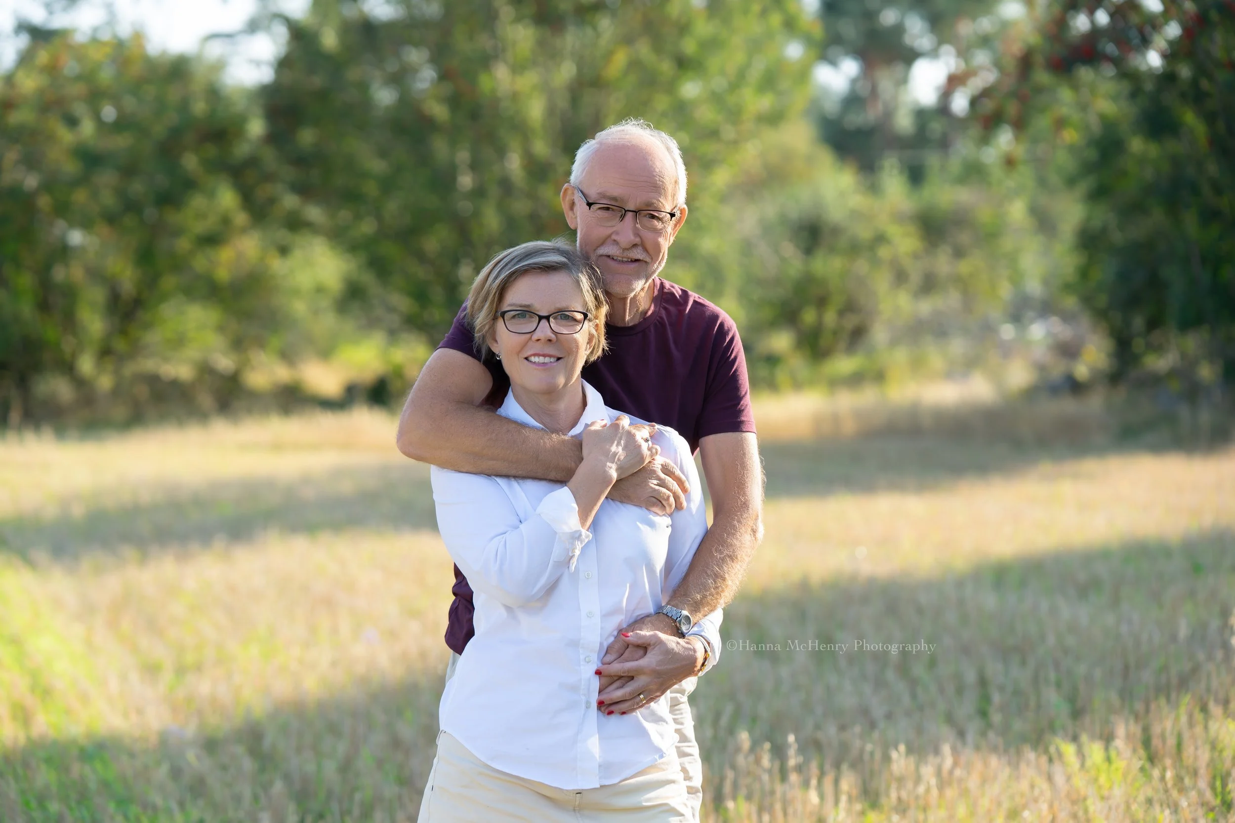 An older man and woman smiling and embracing outdoors in a grassy field with trees in the background.