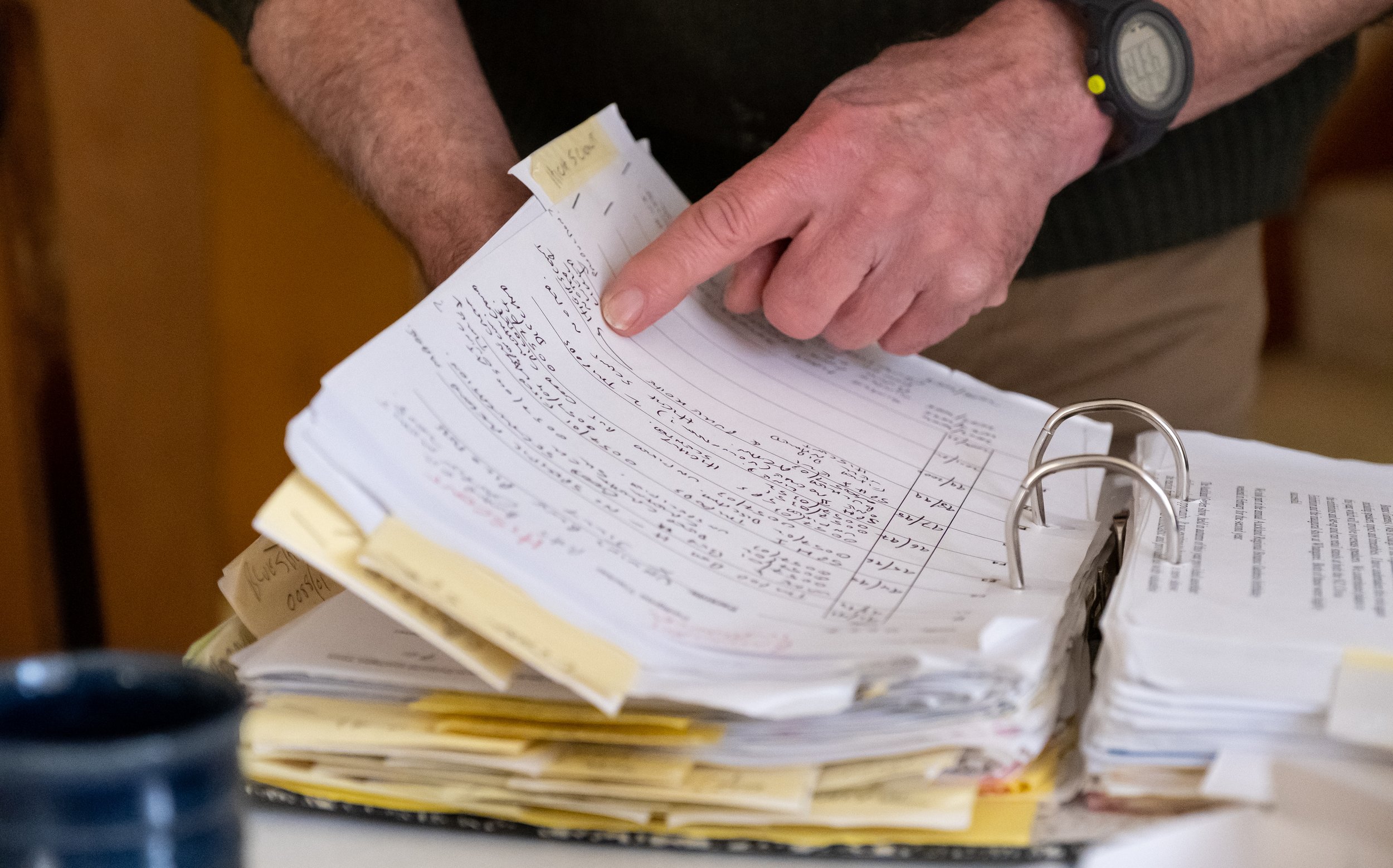Close-up of a person's hand pointing to a page of handwritten notes in a binder filled with similar documents.