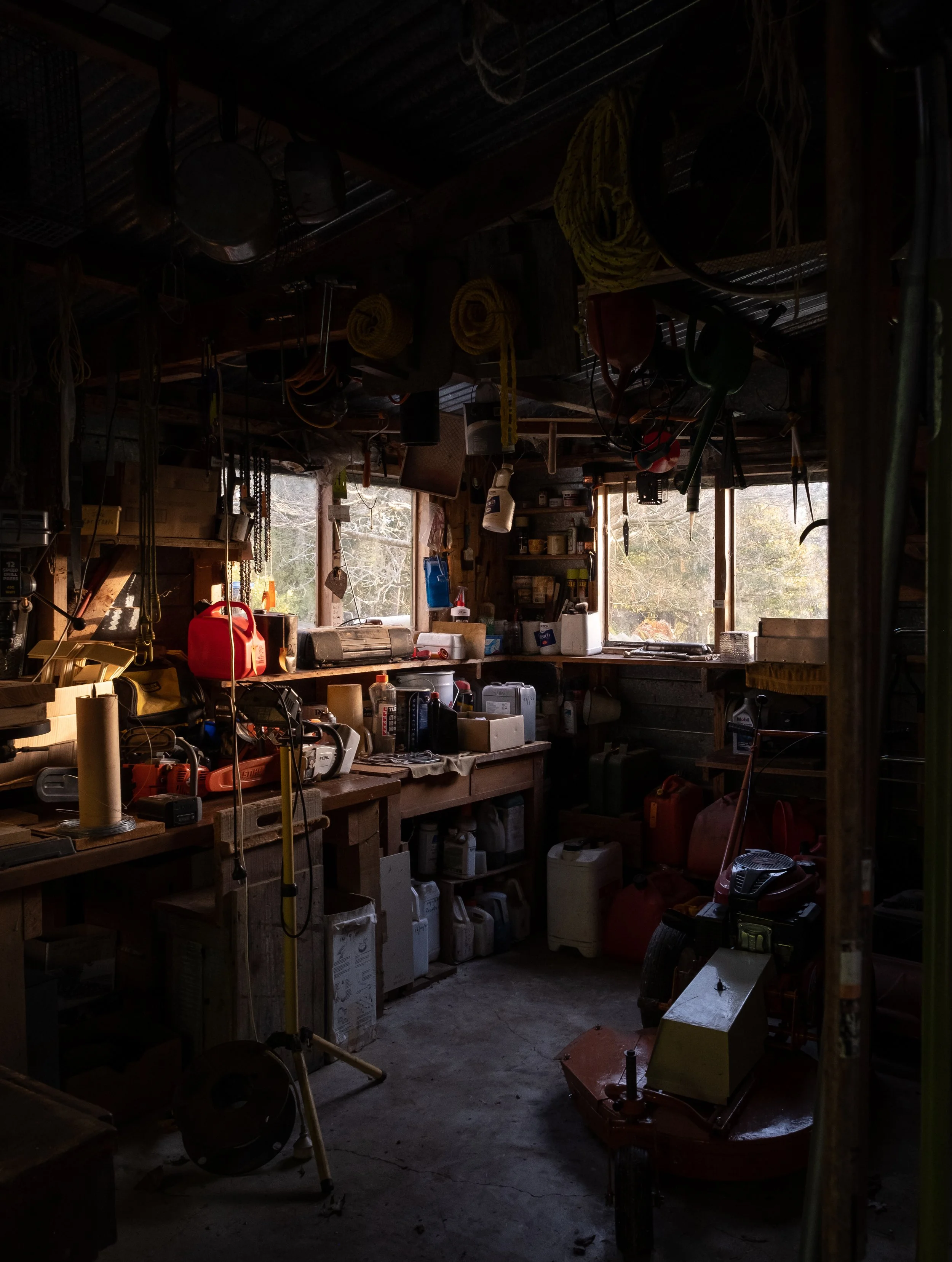 A dimly lit workshop or shed with various tools, equipment, and supplies hanging from the ceiling and placed on shelves and workbenches. There are windows allowing natural light to enter.