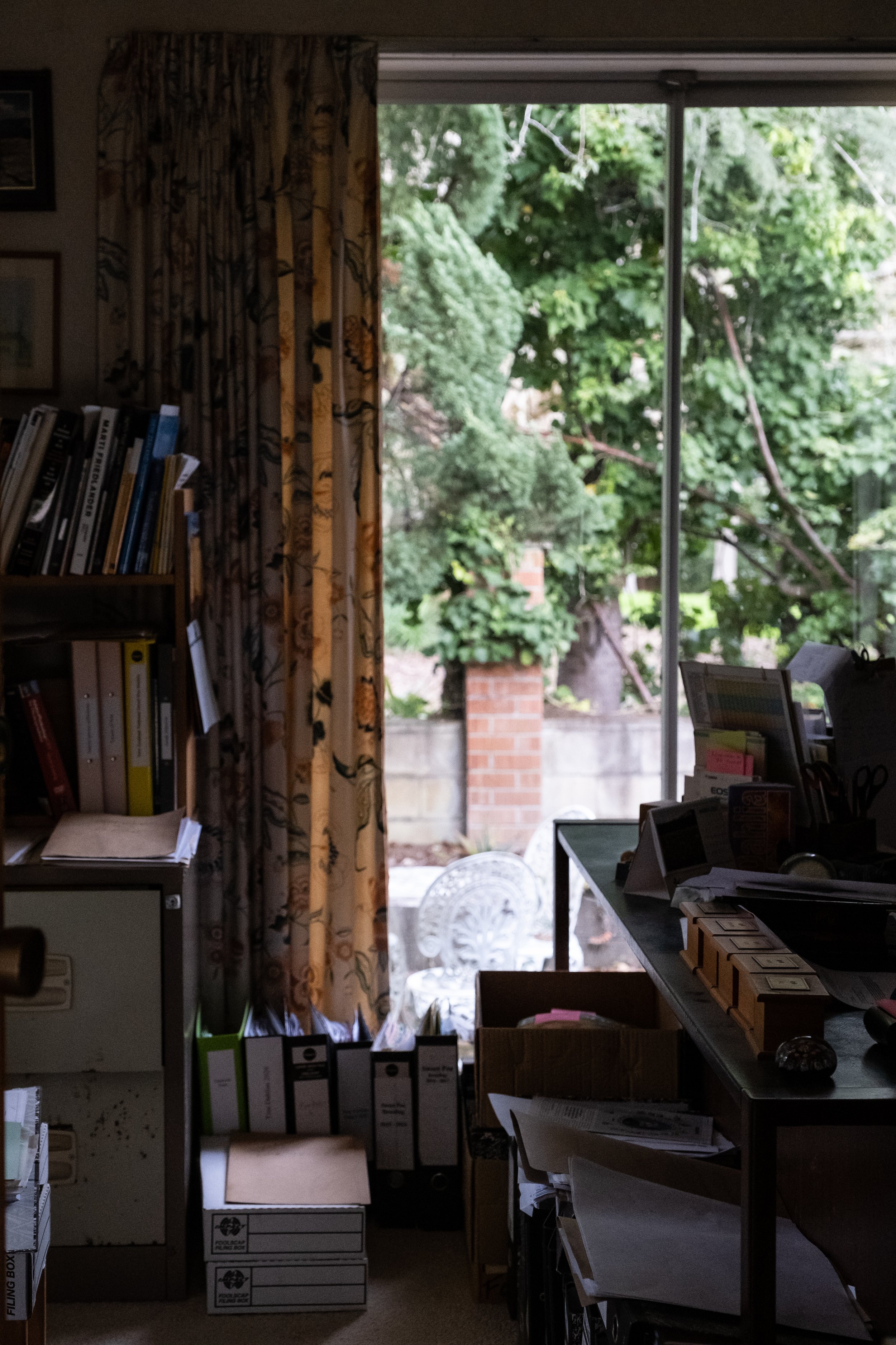 A cluttered office or workspace with shelves filled with binders and files, a desk with papers, office supplies, and a large window showing green trees outside.