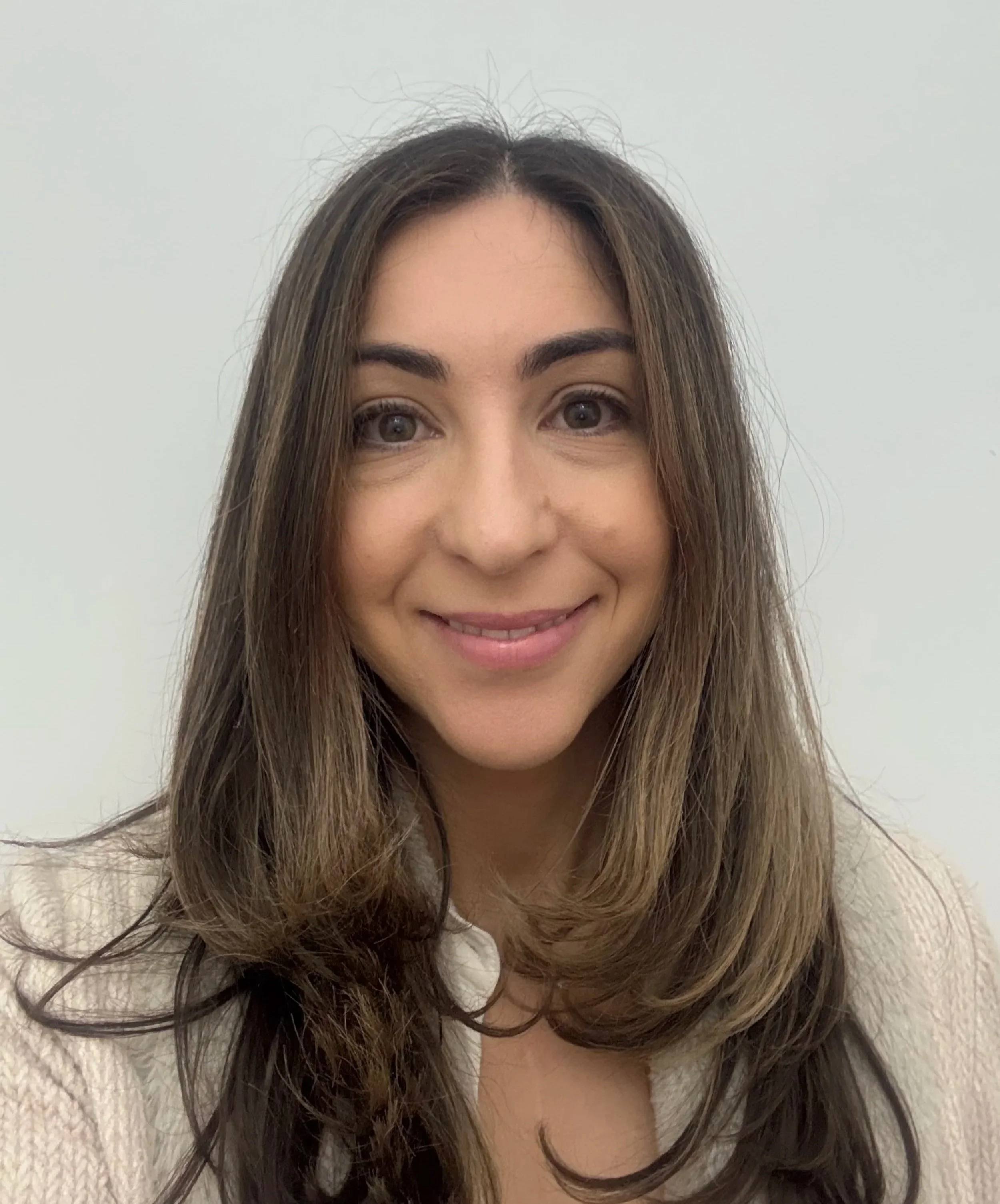 Close-up photo of a woman smiling with long, wavy brown hair and natural makeup, wearing a beige sweater against a plain white wall.