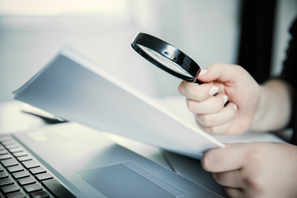 Person holding a magnifying glass and examining documents near a laptop.