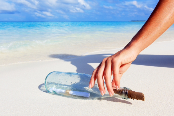 A person placing a message in a bottle on a sandy beach with blue ocean and sky in the background.