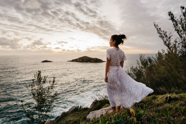 A woman in a white dress with black polka dots standing on a grassy cliff overlooking the ocean at sunset.