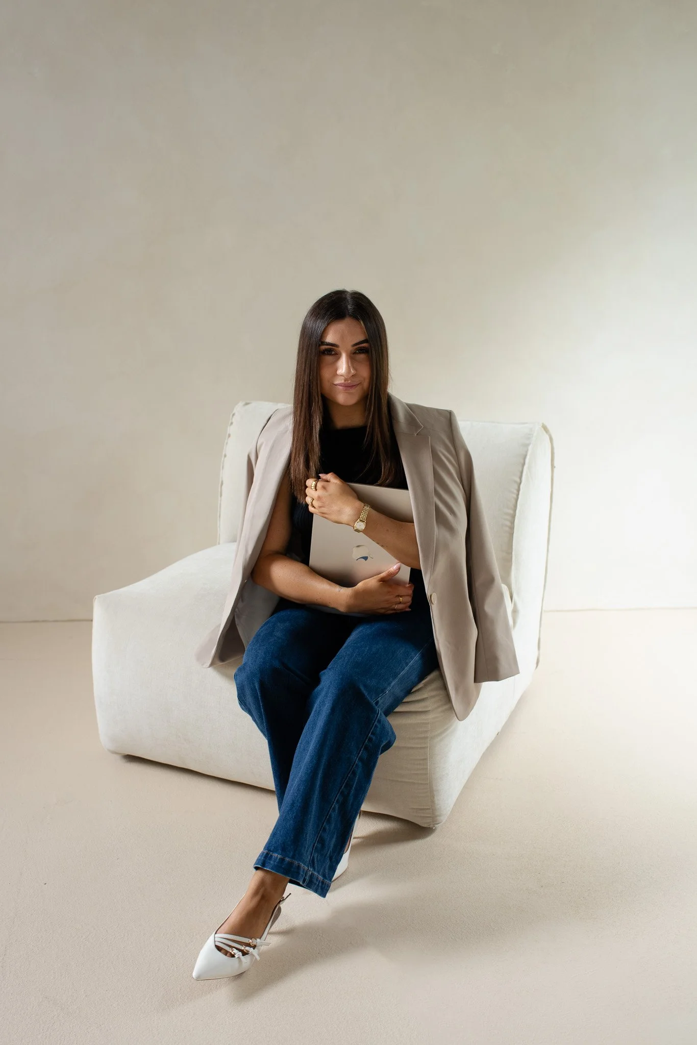 A woman sitting on a cream-colored armchair, holding a notebook or tablet, wearing a black top, blue jeans, white heels, and a beige blazer draped over her shoulders, in a neutral-colored room.