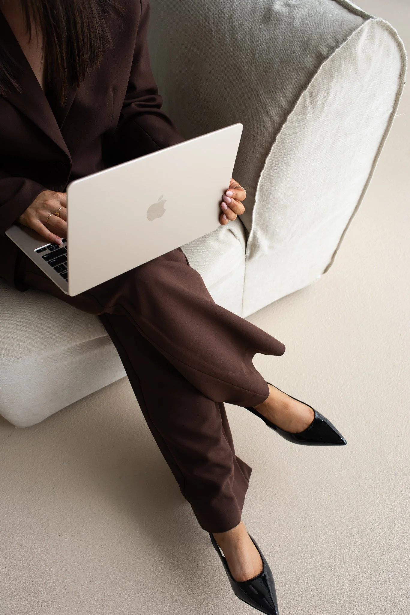 Person sitting on a sofa using a white Apple MacBook laptop. The person is dressed in brown formal pants and is wearing black pointed-toe heels.