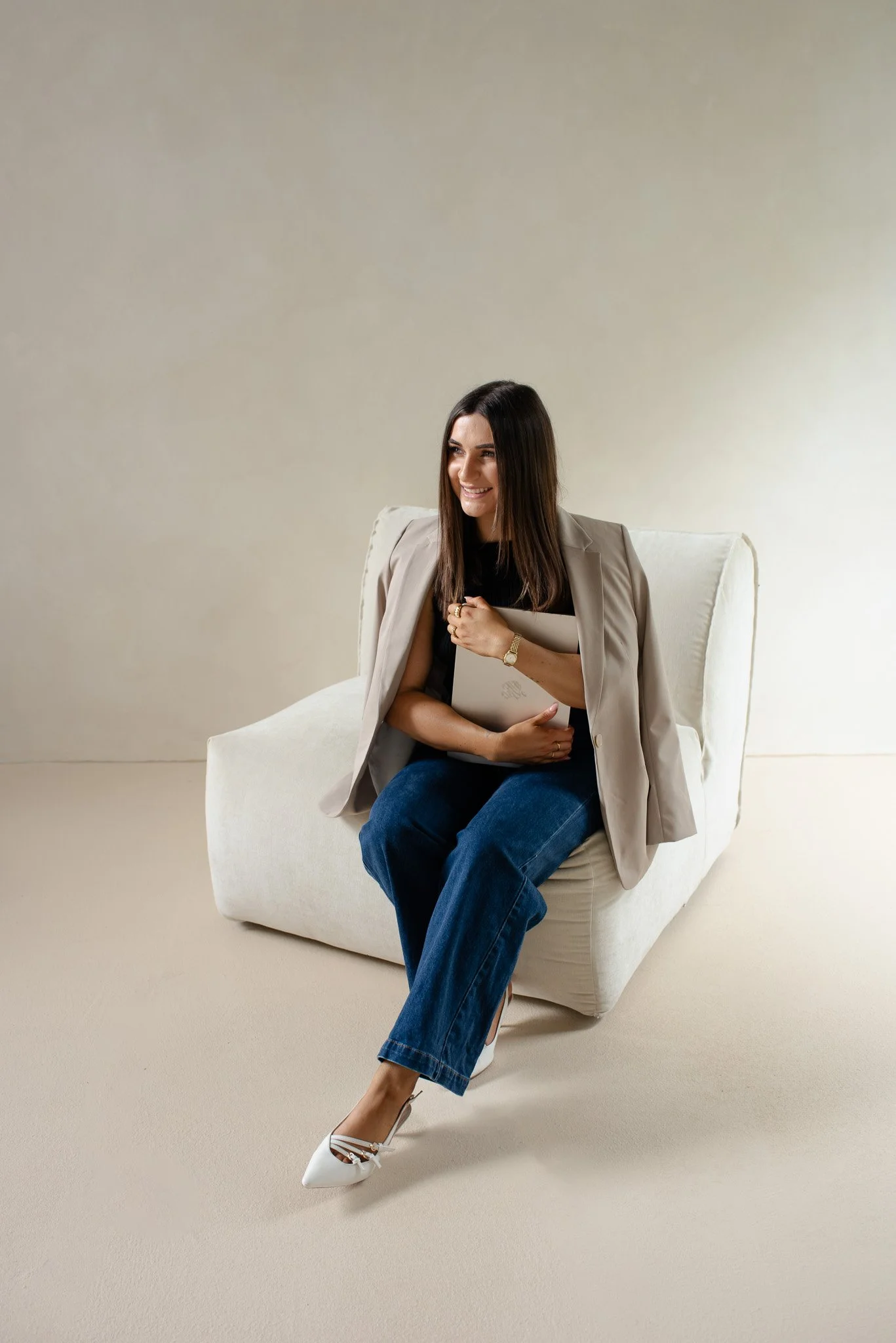 A woman sitting on a light-colored armchair, holding a closed laptop and smiling, dressed in a beige blazer, black top, blue jeans, and white heels, against a plain light background.