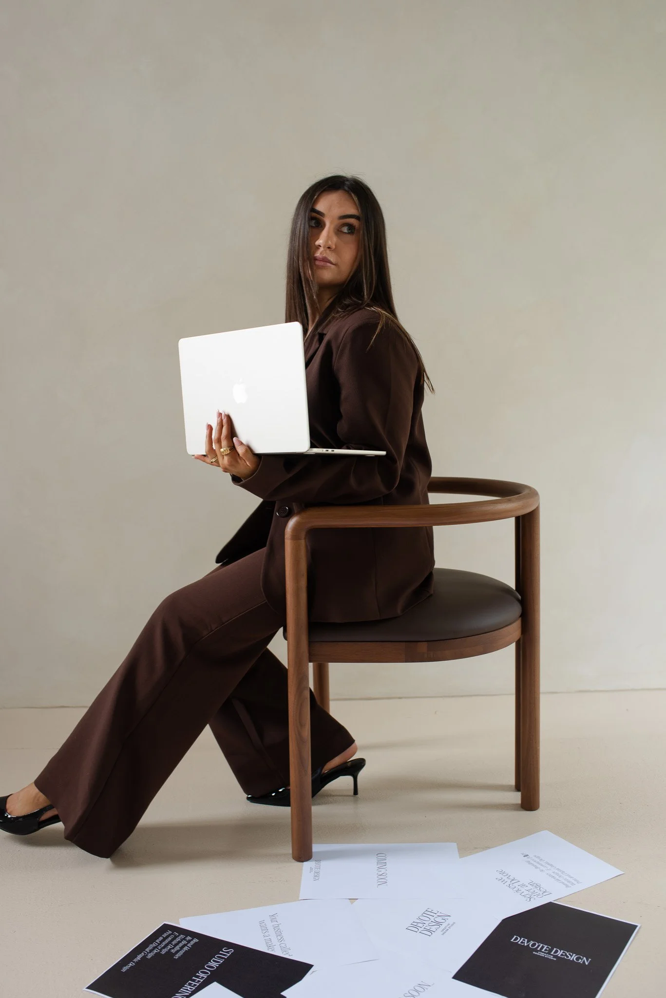 A woman in a brown suit sitting on a wooden chair, holding a silver laptop, with papers scattered on the floor.