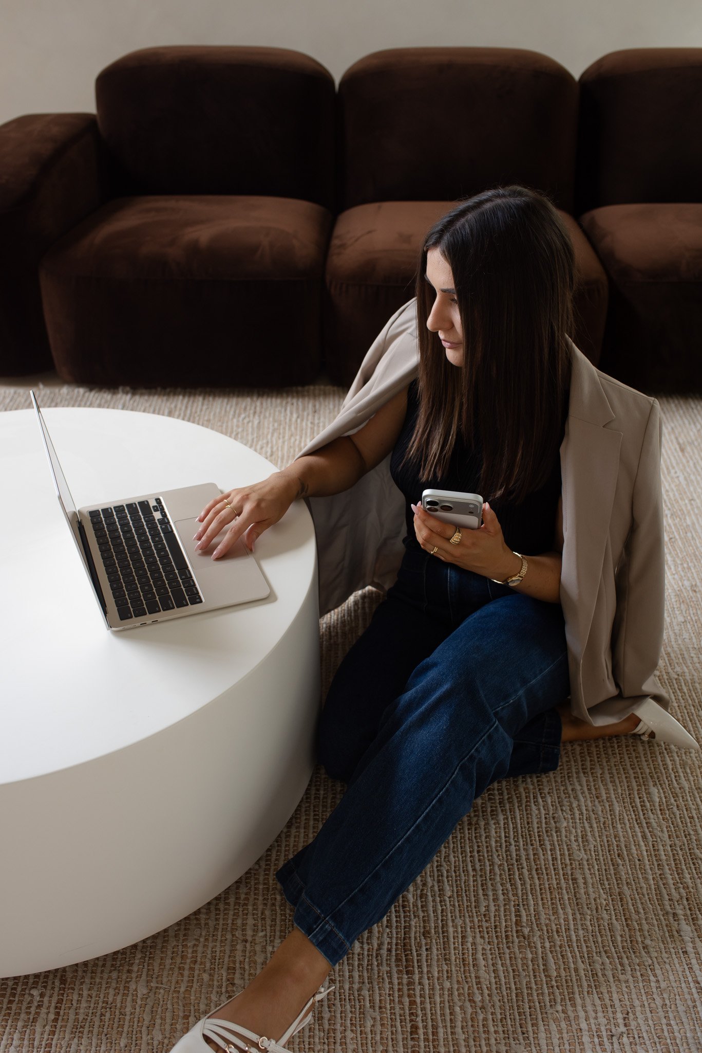 A woman sitting on the floor with a laptop on a white circular table, holding her phone, in a room with a brown couch in the background.