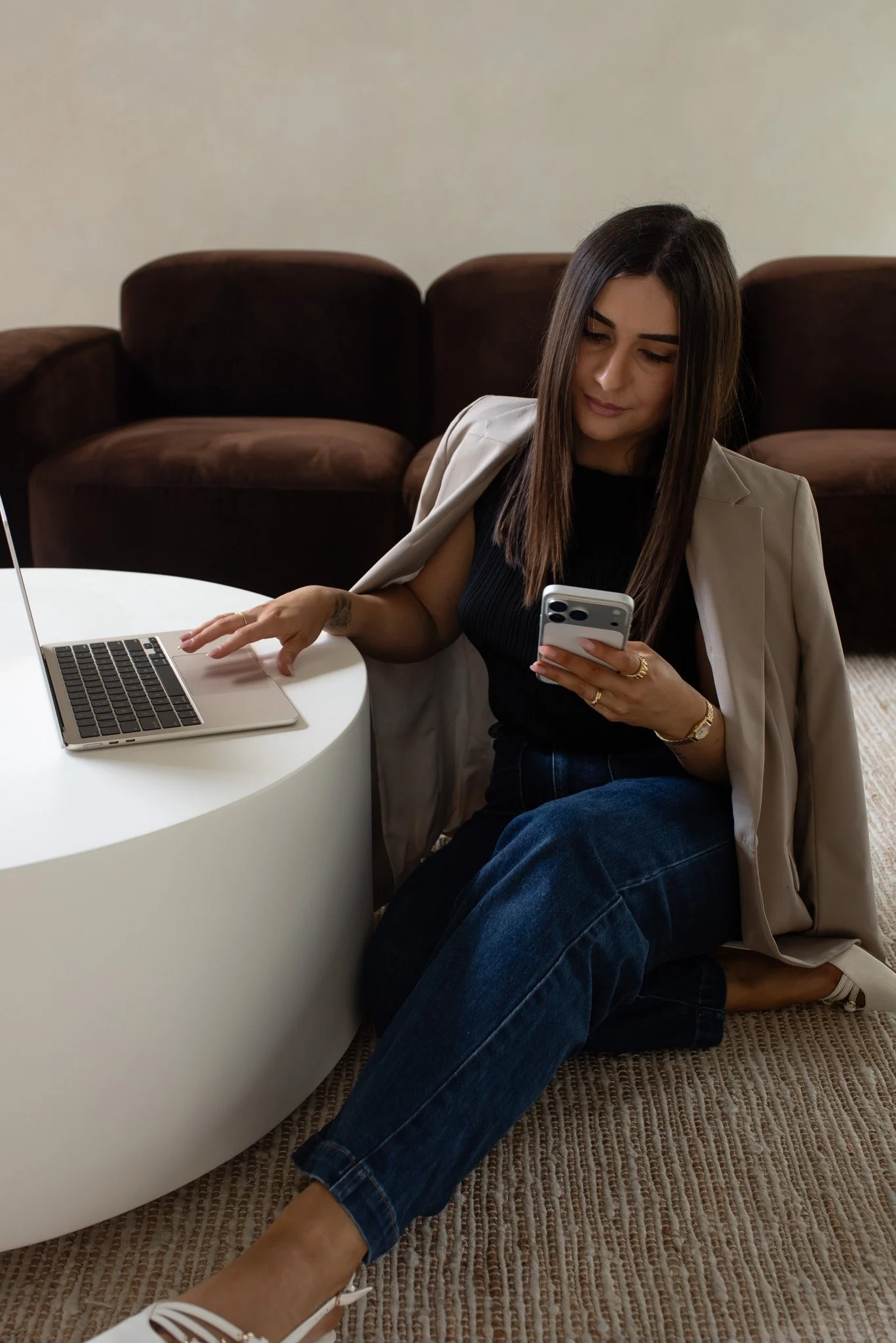 Woman sitting on the floor, using a phone and positioned next to a white coffee table with a laptop on it, with a brown sofa in the background.