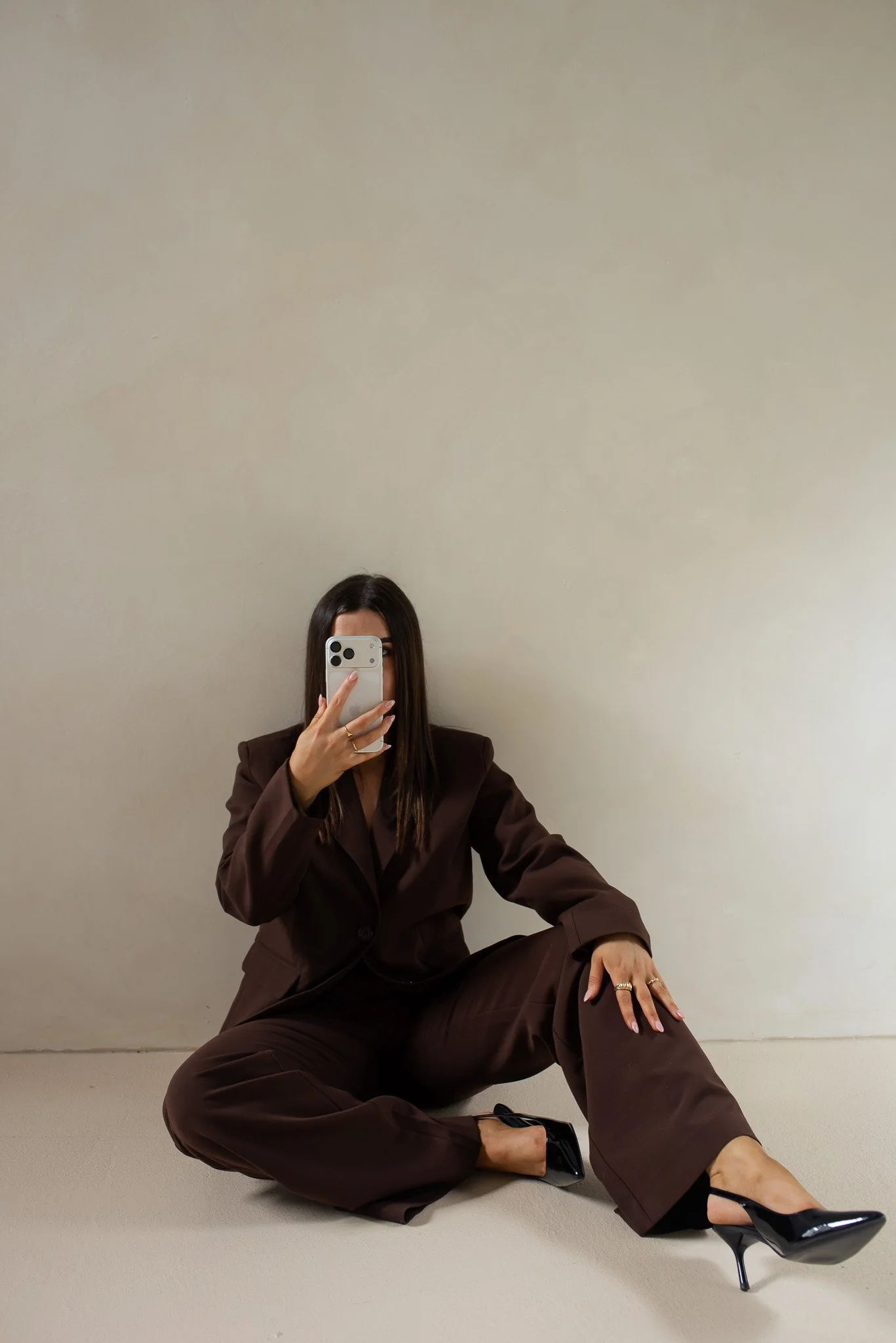 A woman in a brown suit and black high heels sitting on the floor, taking a mirror selfie with a smartphone in a plain room with a beige wall.