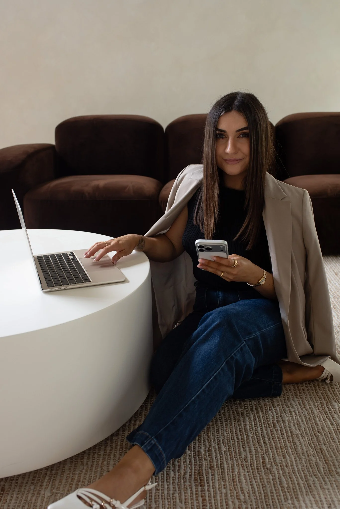 A woman with long brown hair sitting on the floor near a white round table, holding a smartphone, with a laptop on the table, and a brown sofa in the background.