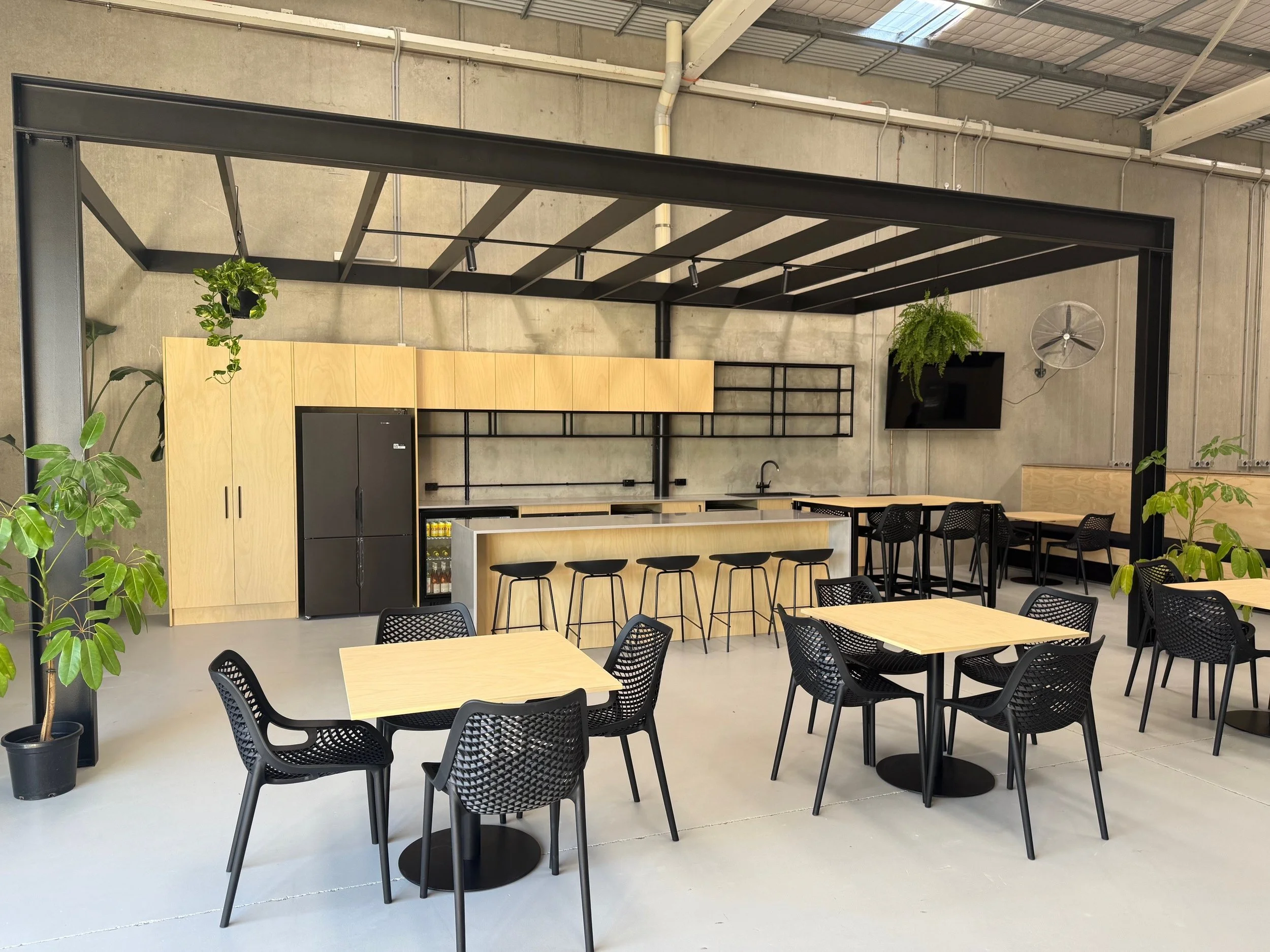 Modern cafeteria with light wood tables, black chairs, industrial ceiling, concrete walls, and green plants.
