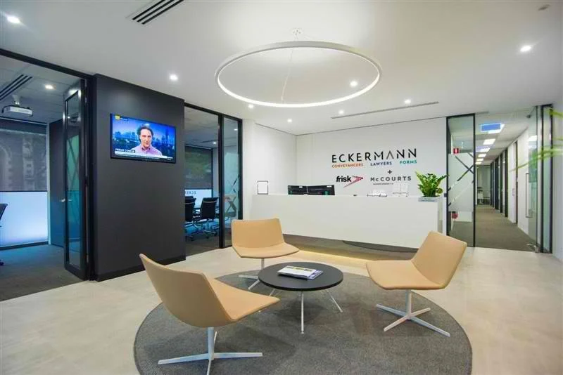 Modern office lobby with a reception desk, a circular light fixture, four beige chairs arranged around a small black table on a gray rug, a wall-mounted TV showing a news broadcast, and glass walls leading to meeting rooms. The company's logo, 'Eckermann', and legal firm names, are displayed behind the reception desk.