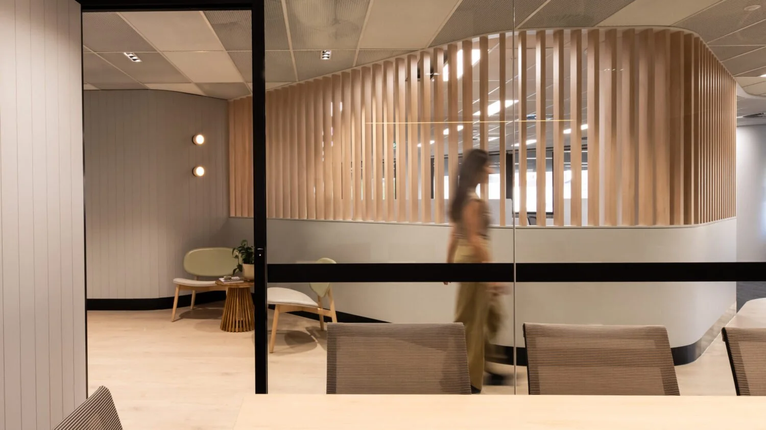 Interior of a modern office space with a woman's silhouette walking past a wooden slatted partition. There are chairs, a small table, a plant, and wall-mounted lights visible, with neutral color tones and a contemporary design.