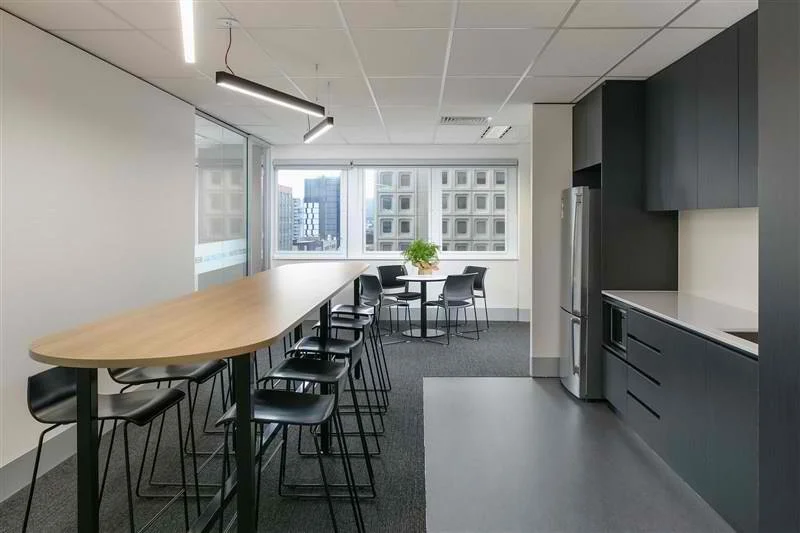Modern office kitchen and communal area with a high dining table and bar stools, a round table with chairs near windows, gray cabinets, and a refrigerator.