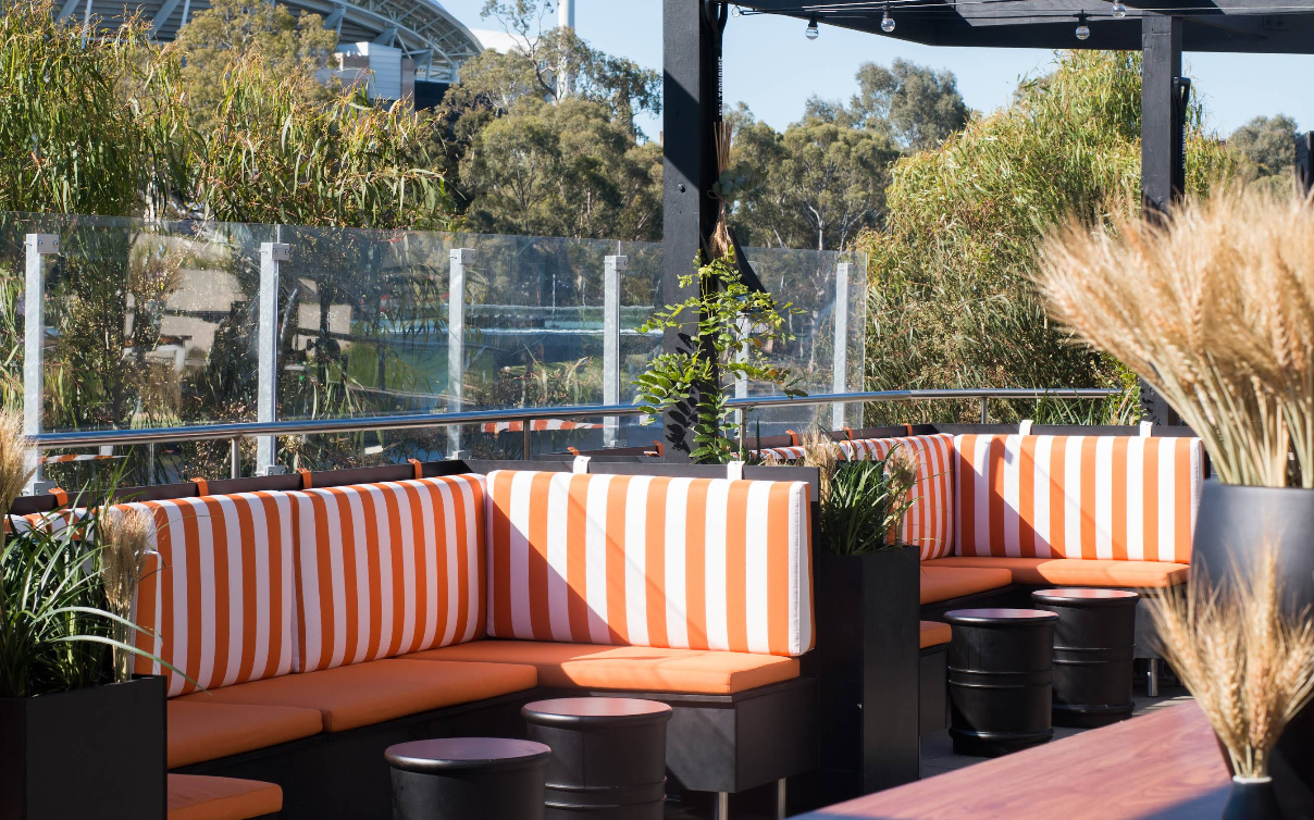 Outdoor seating area with orange and white striped cushions, black tables, and potted plants, with glass railing and trees in the background.