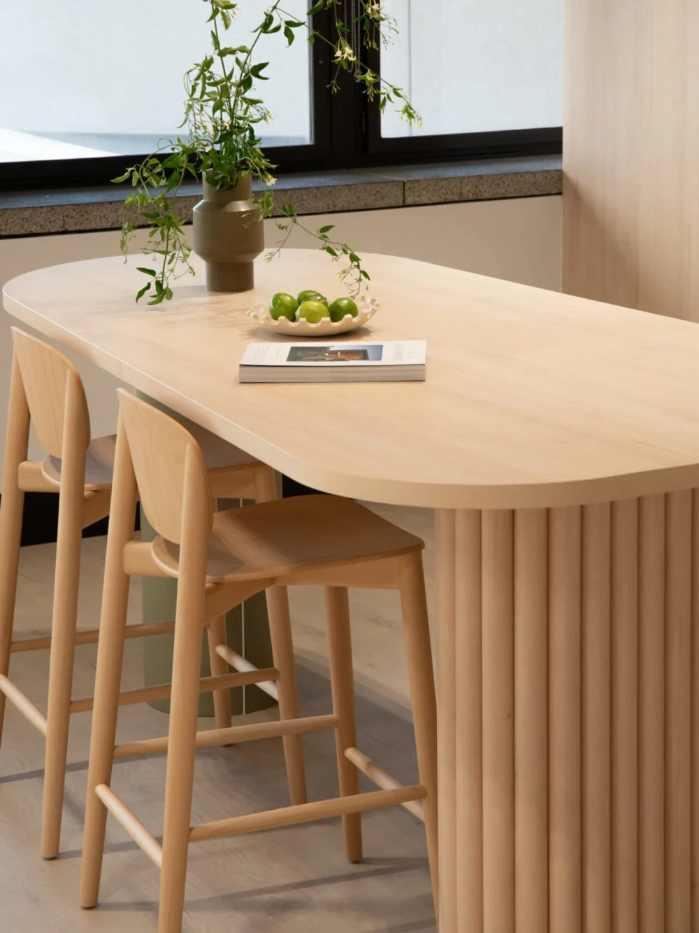 A light wood dining table with three matching wooden chairs, a ceramic vase with green leafy plant, a small ceramic bowl with green apples, and a magazine, with a window in the background.