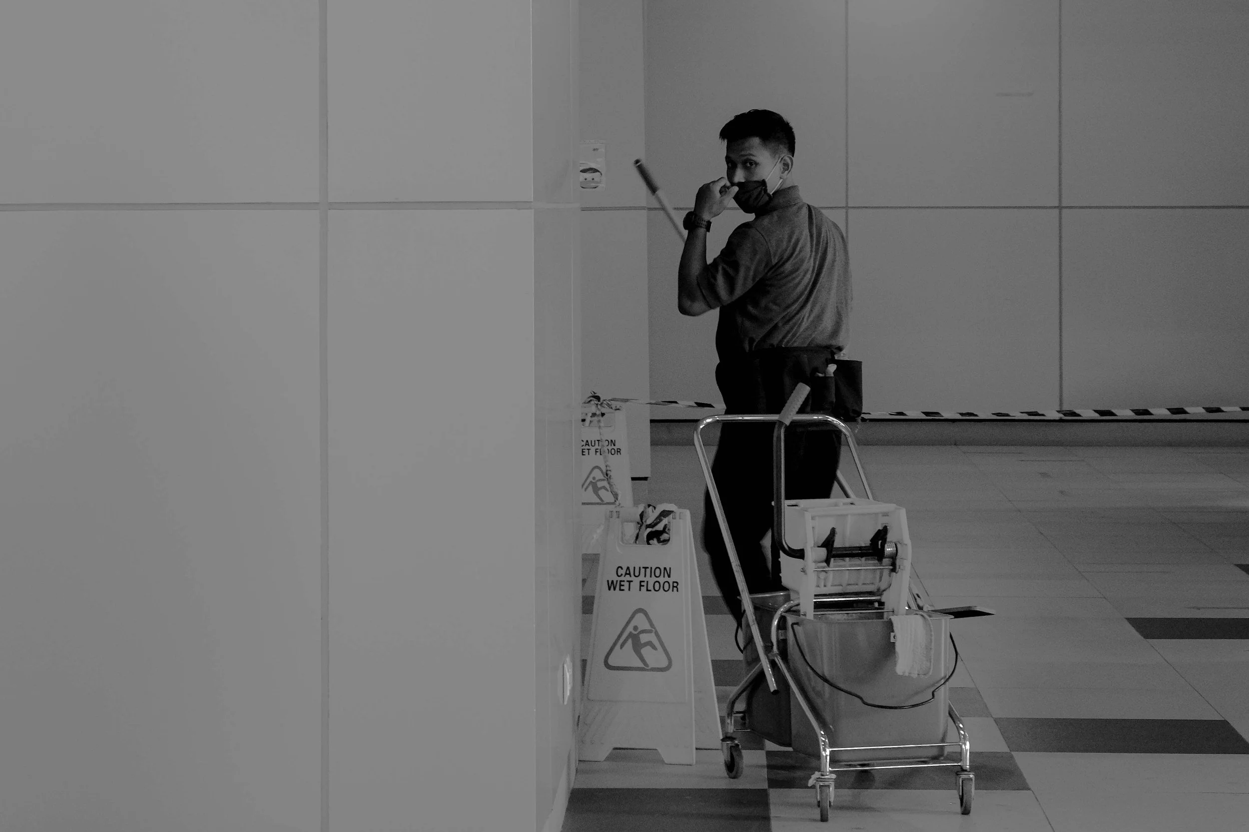A man with short dark hair wearing a face mask, standing in an airport or similar facility, holds a smartphone to his face. He is surrounded by bilingual caution signs warning of a wet floor, and has a luggage trolley nearby.