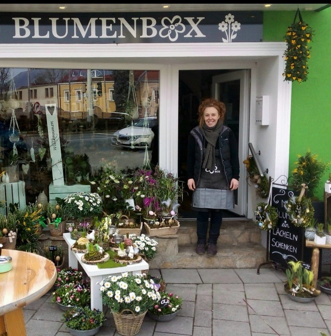 Eine Frau steht vor einem Blumengeschäft mit dem Schild 'Blumenbox'. Im Schaufenster sind verschiedene Pflanzen und Dekorationen zu sehen. Vor dem Geschäft sind viele Blumen und Ostereier zum Verkauf ausgestellt.