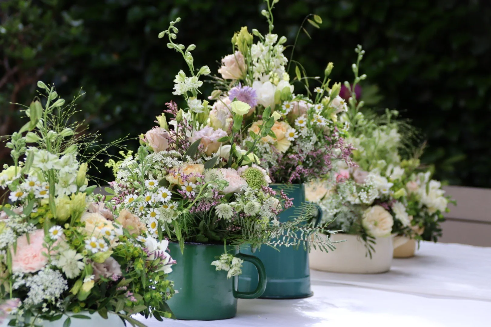 Bunte Blumenarrangements in Tassen auf einem weißen Tisch im Freien.