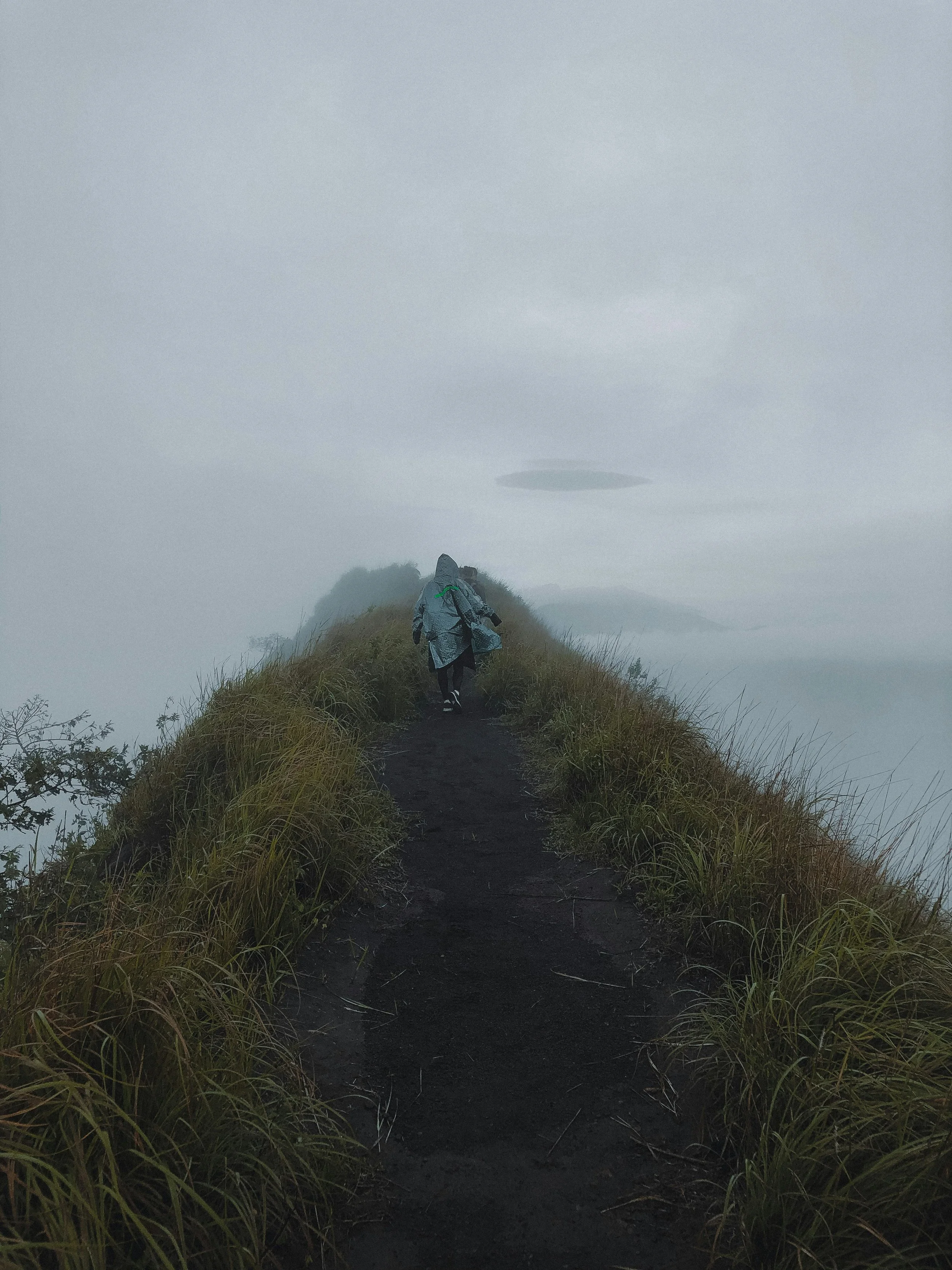 Deux personnes marchent sur un sentier étroit et sombre au sommet d'une colline ou d'une montagne, entourées de brouillard ou de nuages, avec un ciel gris et brumeux.