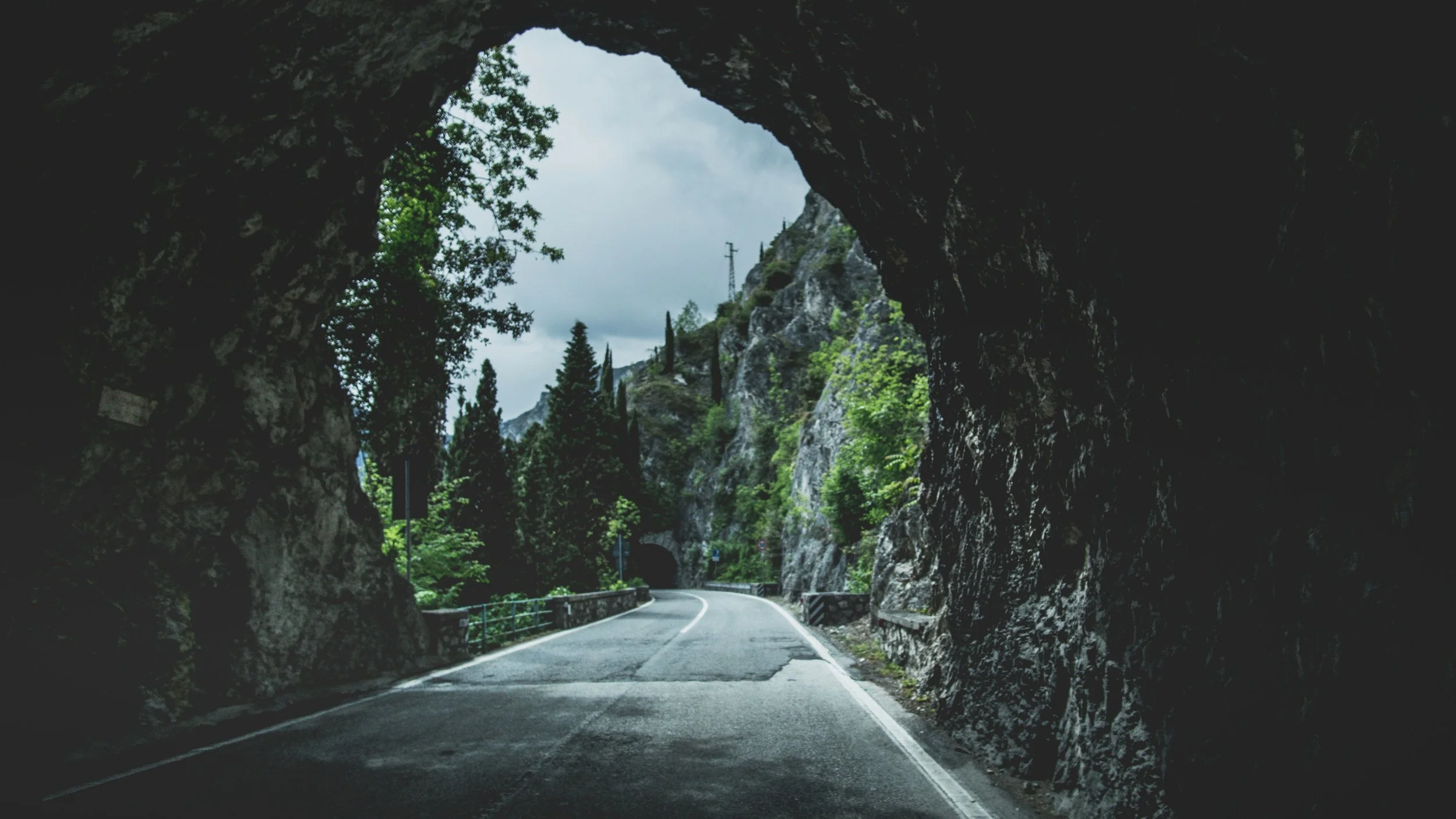 Vue d'une route passant à travers une grotte rocheuse, entourée de montagnes et d'arbres verts, avec un ciel nuageux.