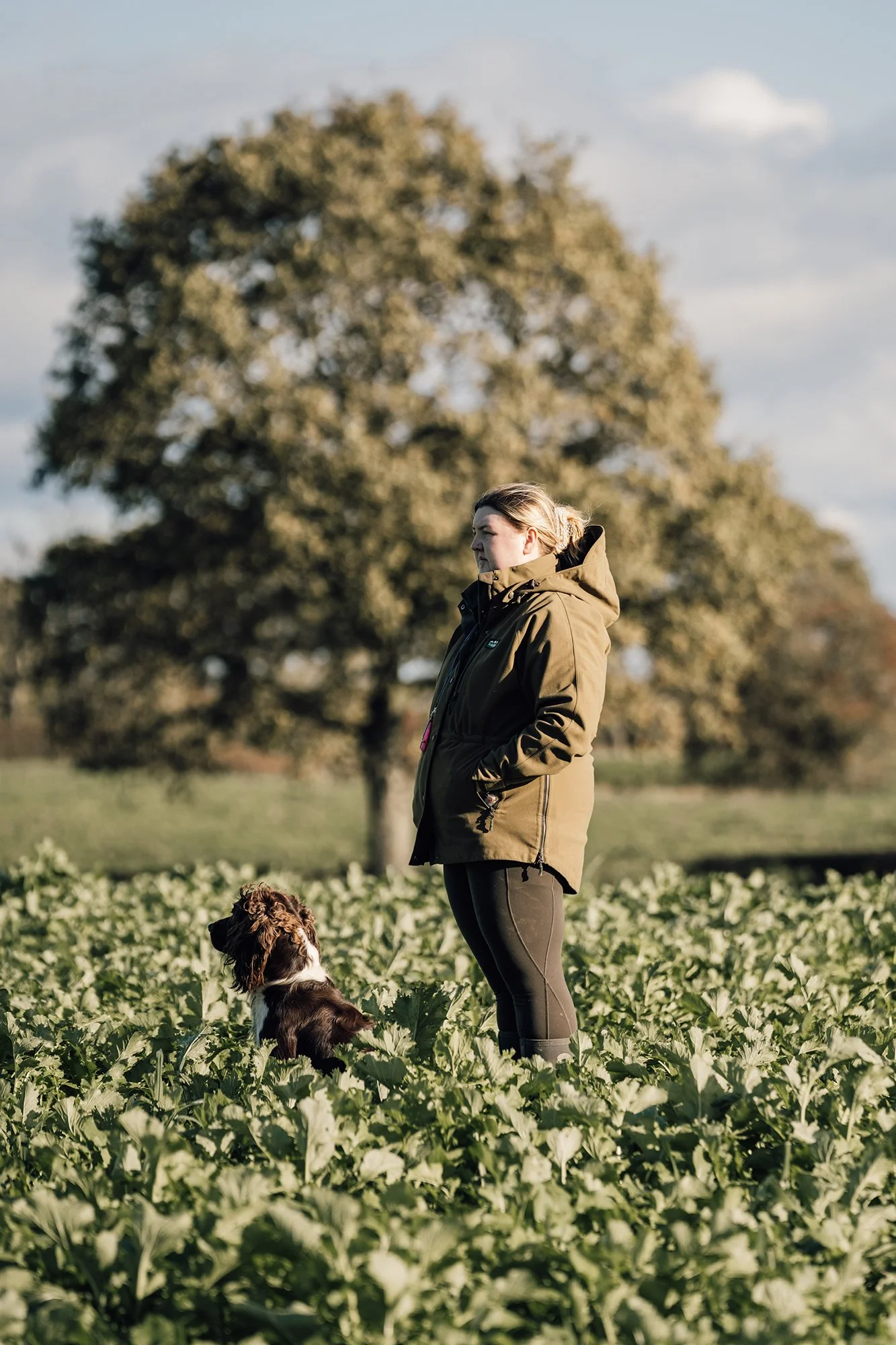 A woman standing in a field of green plants, with a large tree in the background, wearing a brown jacket and black pants, and a dog sitting beside her.