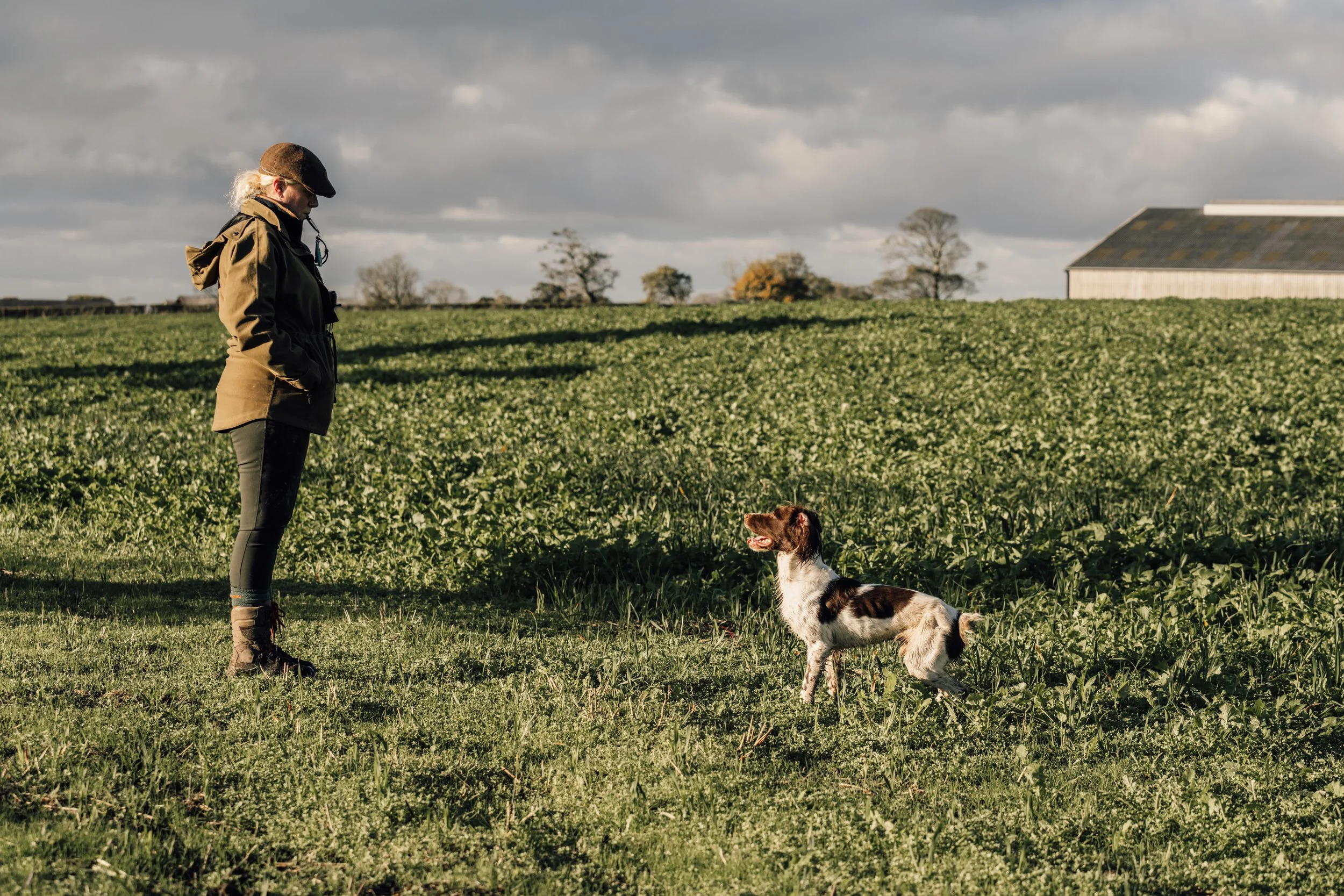 A person in outdoor gear standing in a grassy field with a brown and white dog sitting nearby. The sky is cloudy, and there are trees and a building in the background.