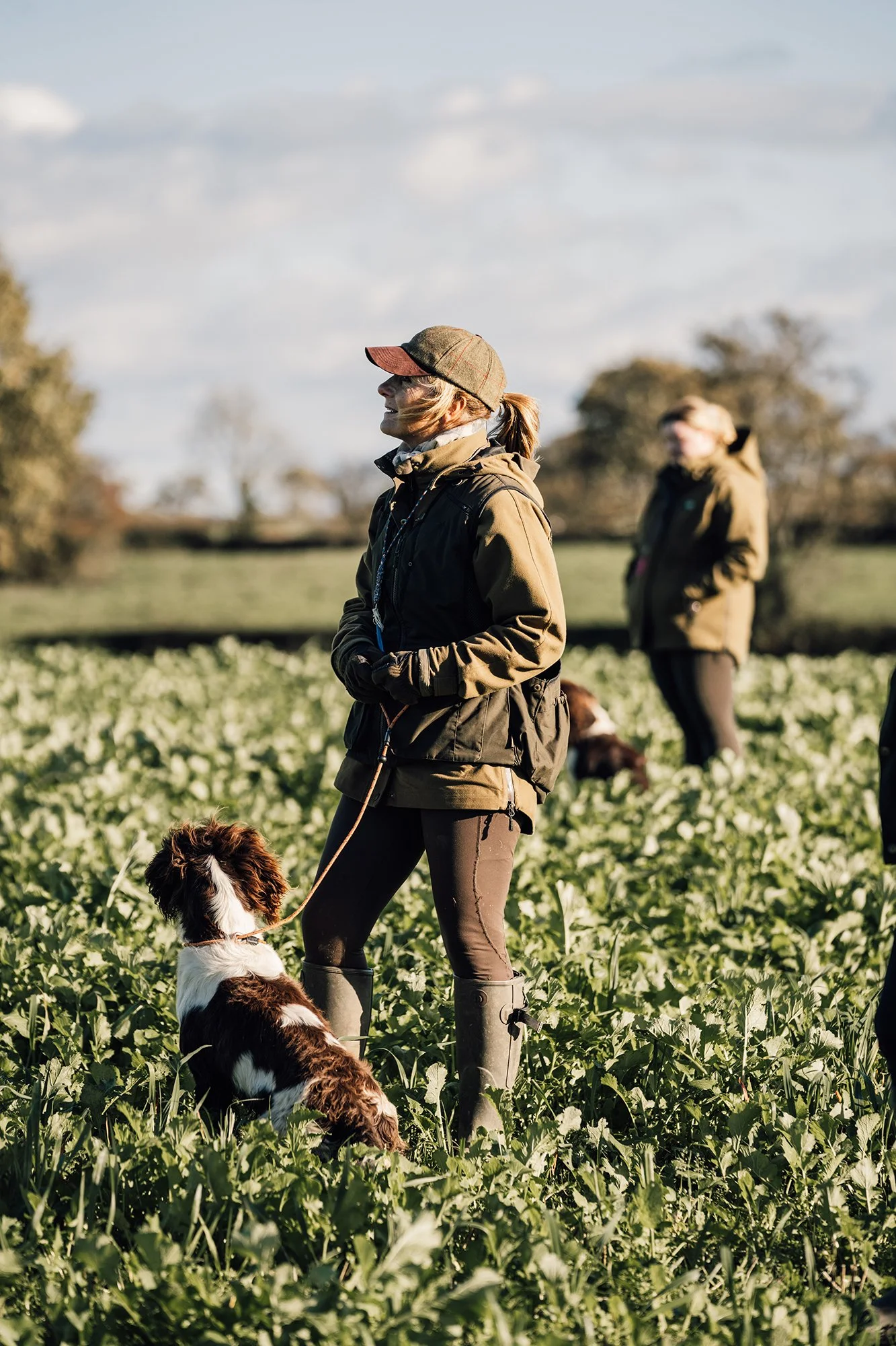People standing in a green field with dogs, dressed in outdoor jackets and boots, enjoying a sunny day.