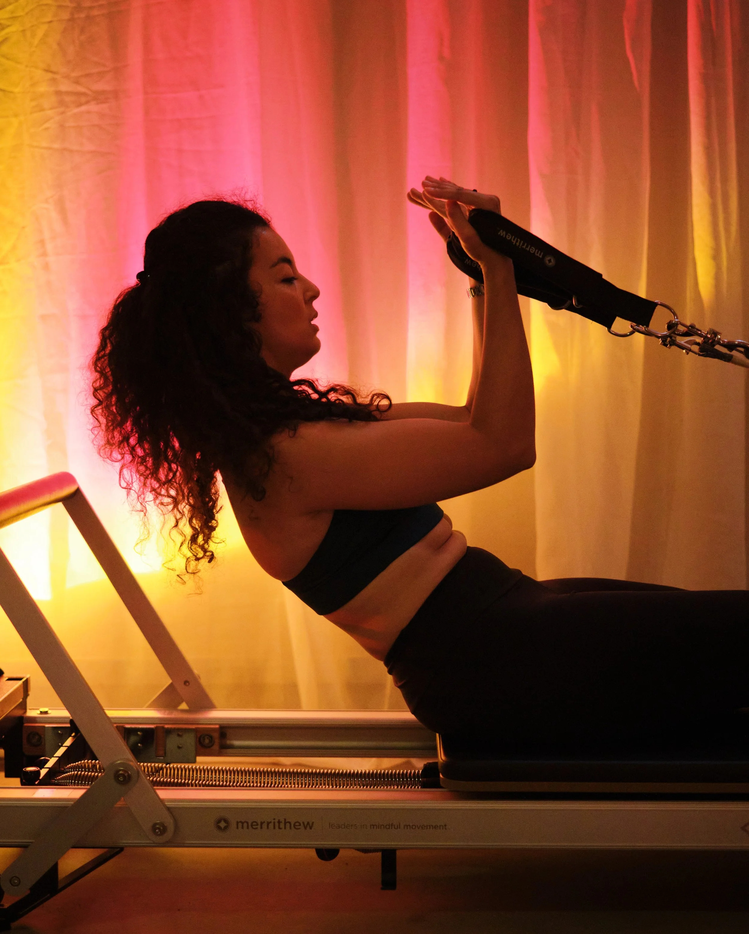 A woman with curly hair exercising on a Pilates reformer machine, performing a core exercise with her arms extended and hands clasped overhead, with a warm pink and yellow lighting background.