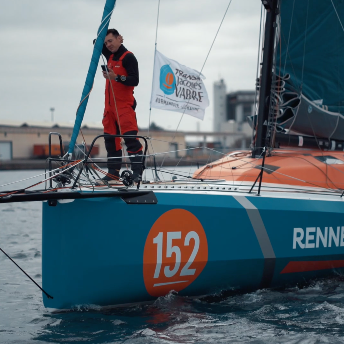 Marina con barca a vela con il numero 152, vela arancione, uomo in tuta rossa e nera che controlla un dispositivo, bandiera con logo e scritte.