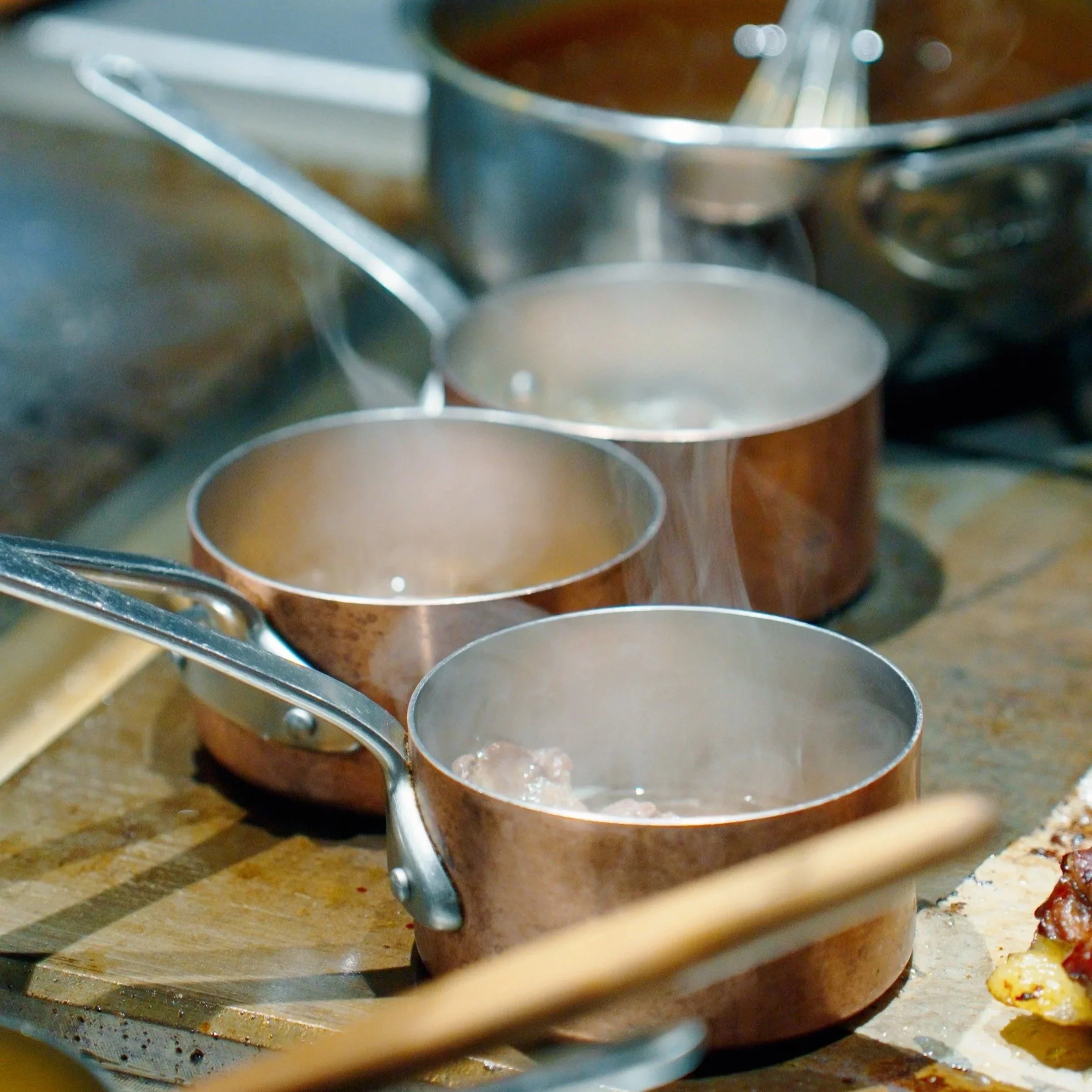 Pentole di rame sul fuoco con vapore che sale, parte di un piano di lavoro in legno, forse in una cucina.